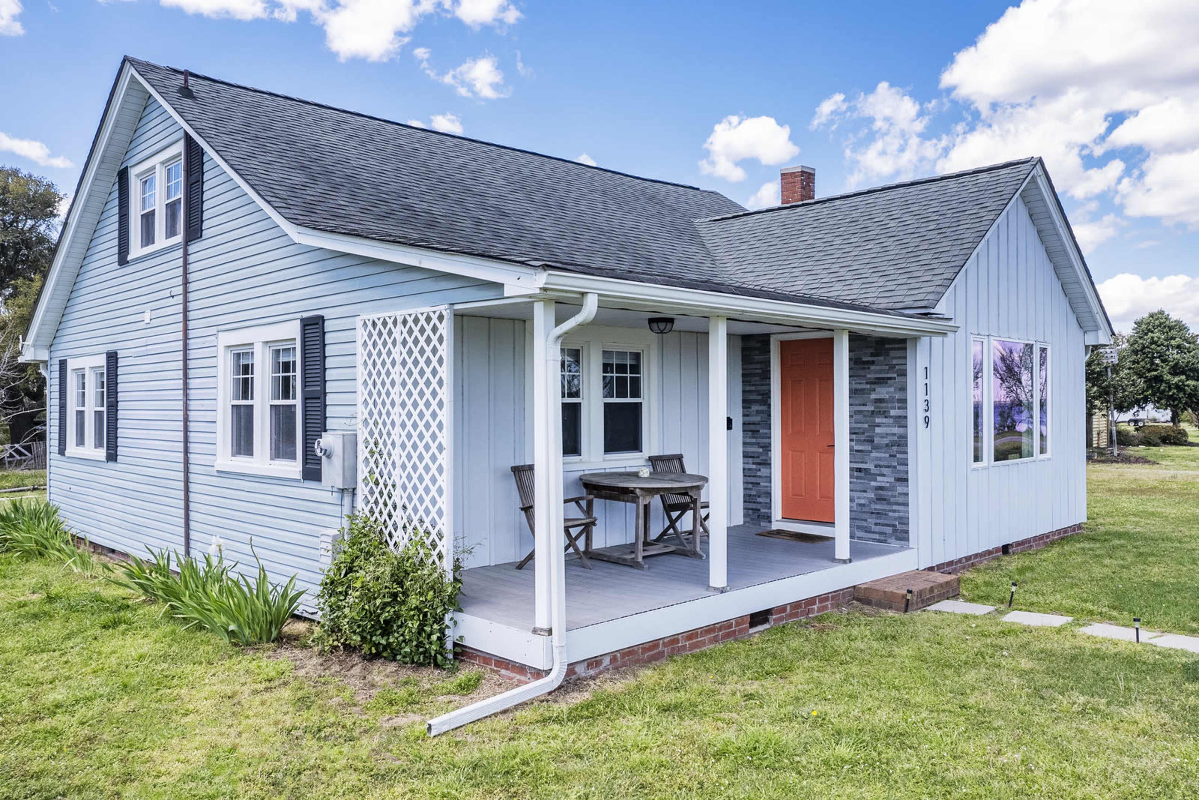 The image shows a light blue, single-story house with a front porch that has two chairs and a small table, surrounded by green grass and a clear blue sky.