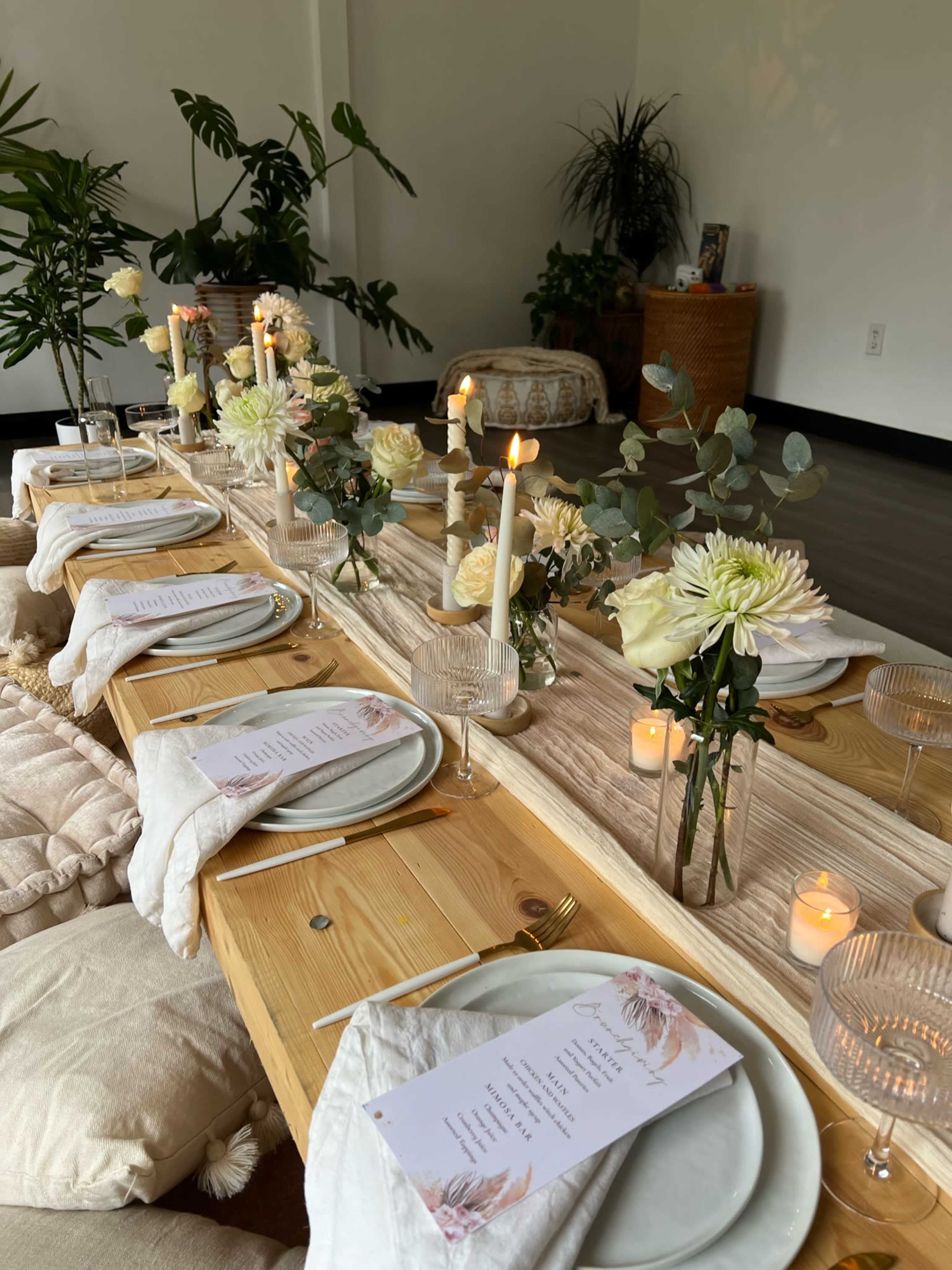 A long wooden table is set for a dinner, adorned with white flowers, candles, and elegant place settings.