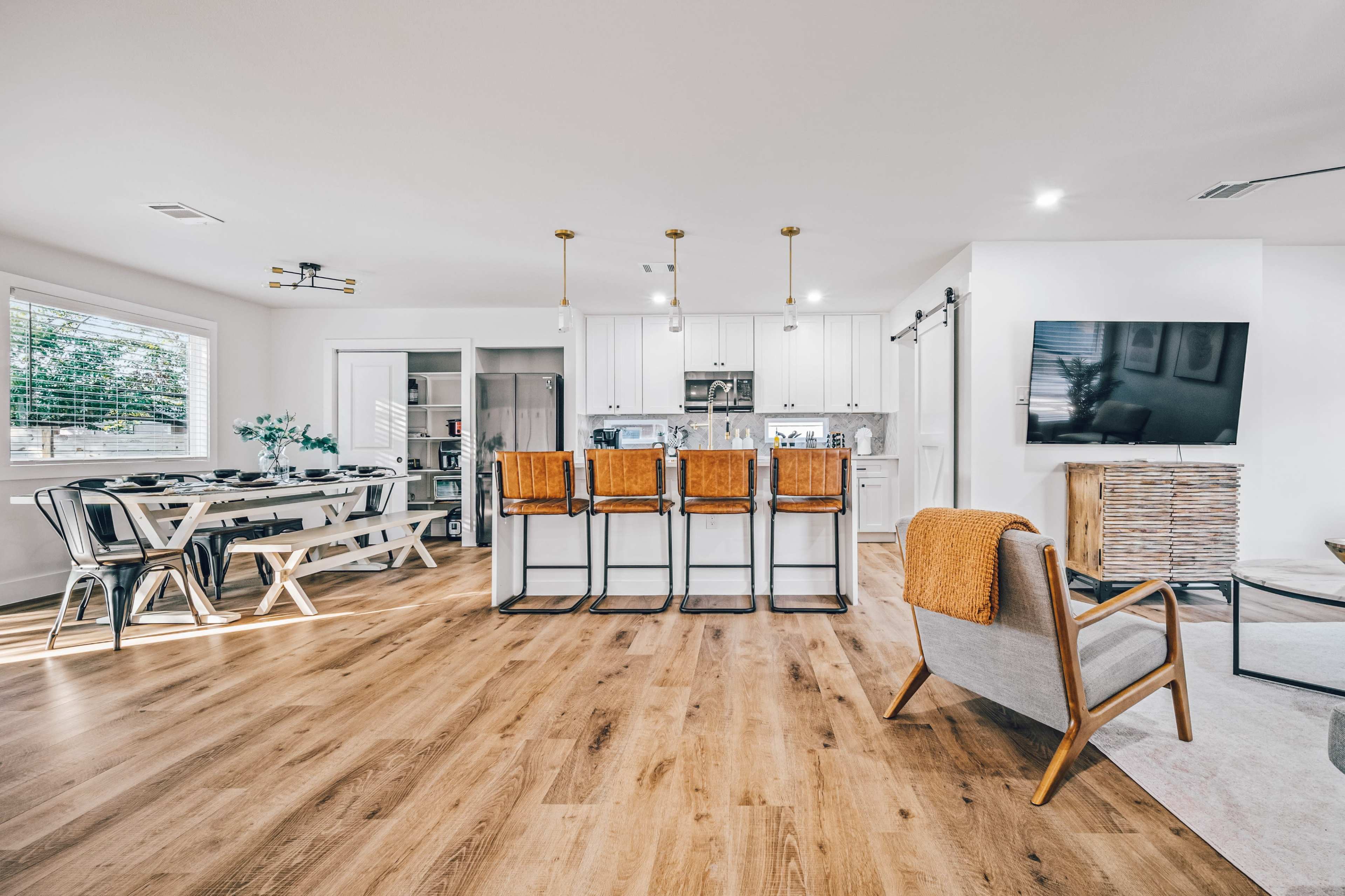 A modern open-concept living space featuring a kitchen island with four bar stools, a dining area with a large table, and a cozy seating arrangement with a television on the wall.
