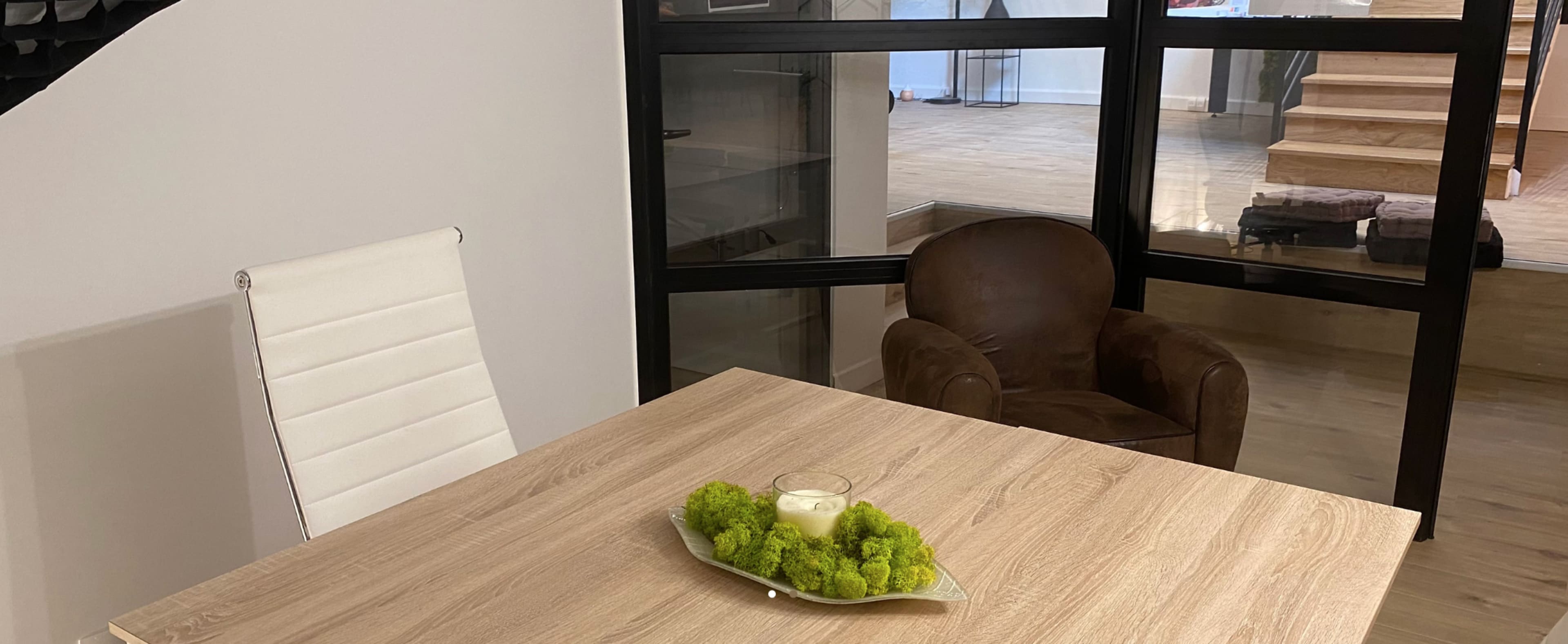 A wooden dining table with a green centerpiece is positioned in front of a glass wall, featuring a white chair and a brown armchair nearby.