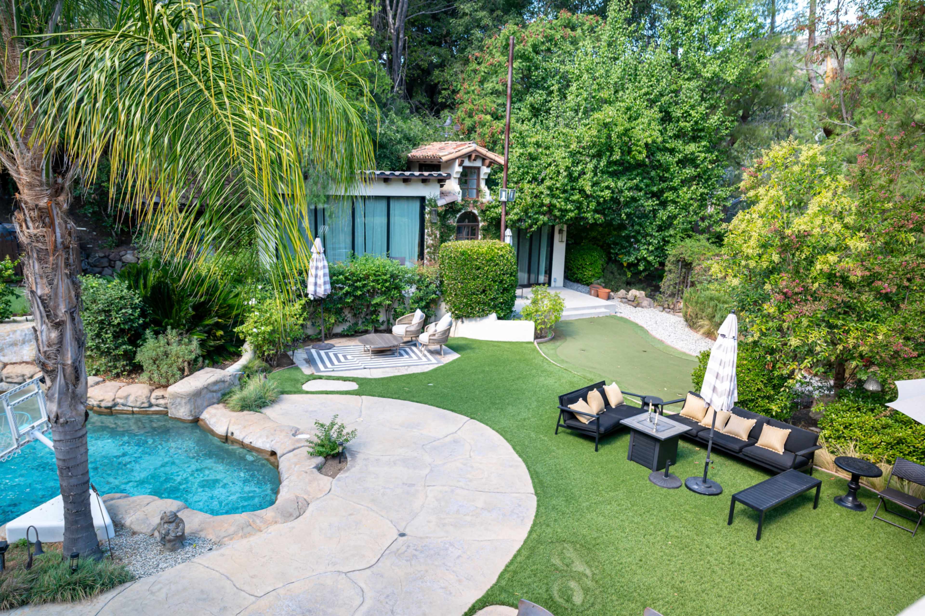 The image shows a landscaped backyard featuring a swimming pool, a patio area with lounge furniture, and a small garden with trees and shrubs.