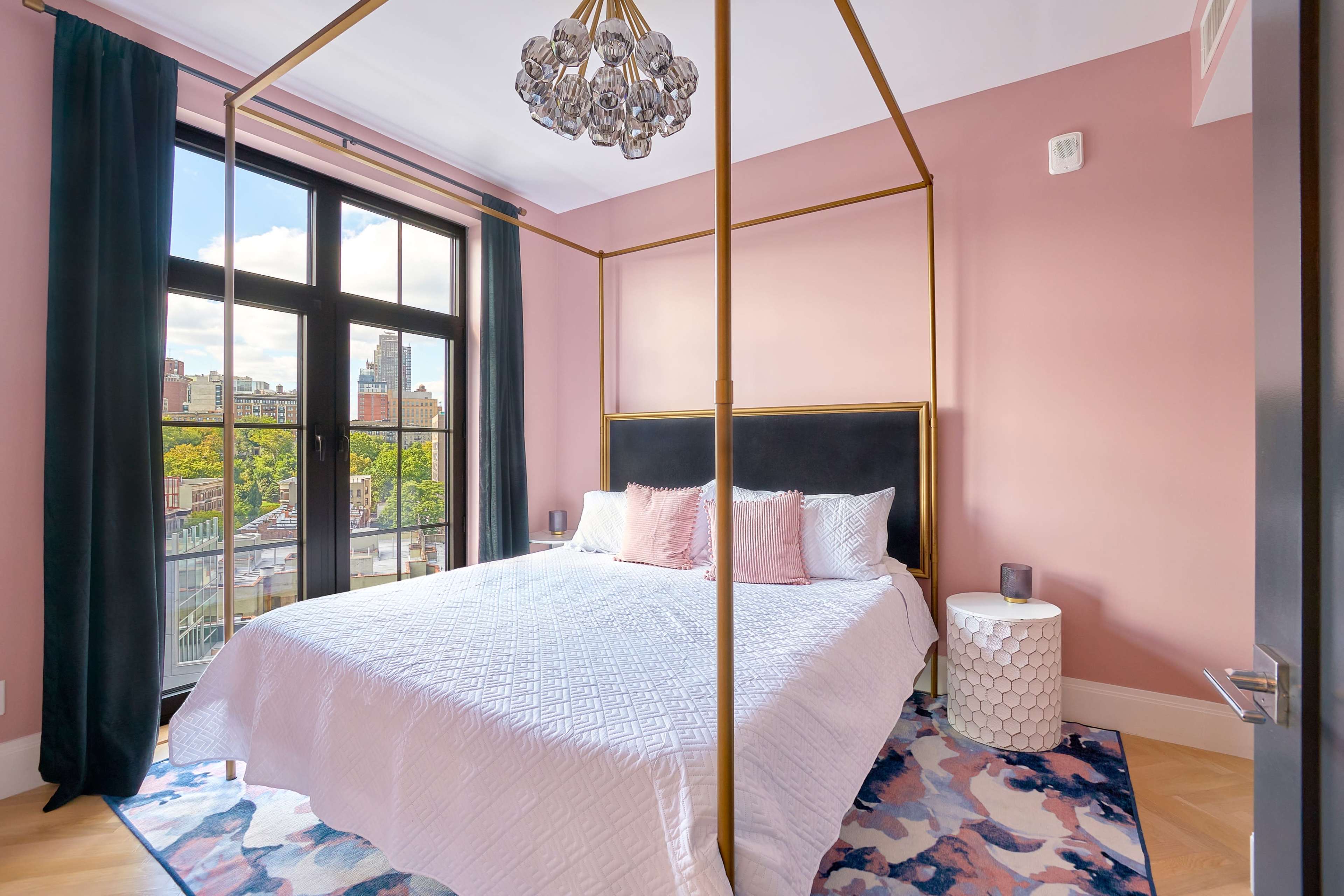A modern bedroom features a four-poster bed with a black headboard, surrounded by pink walls, and large windows offering a view of the city.