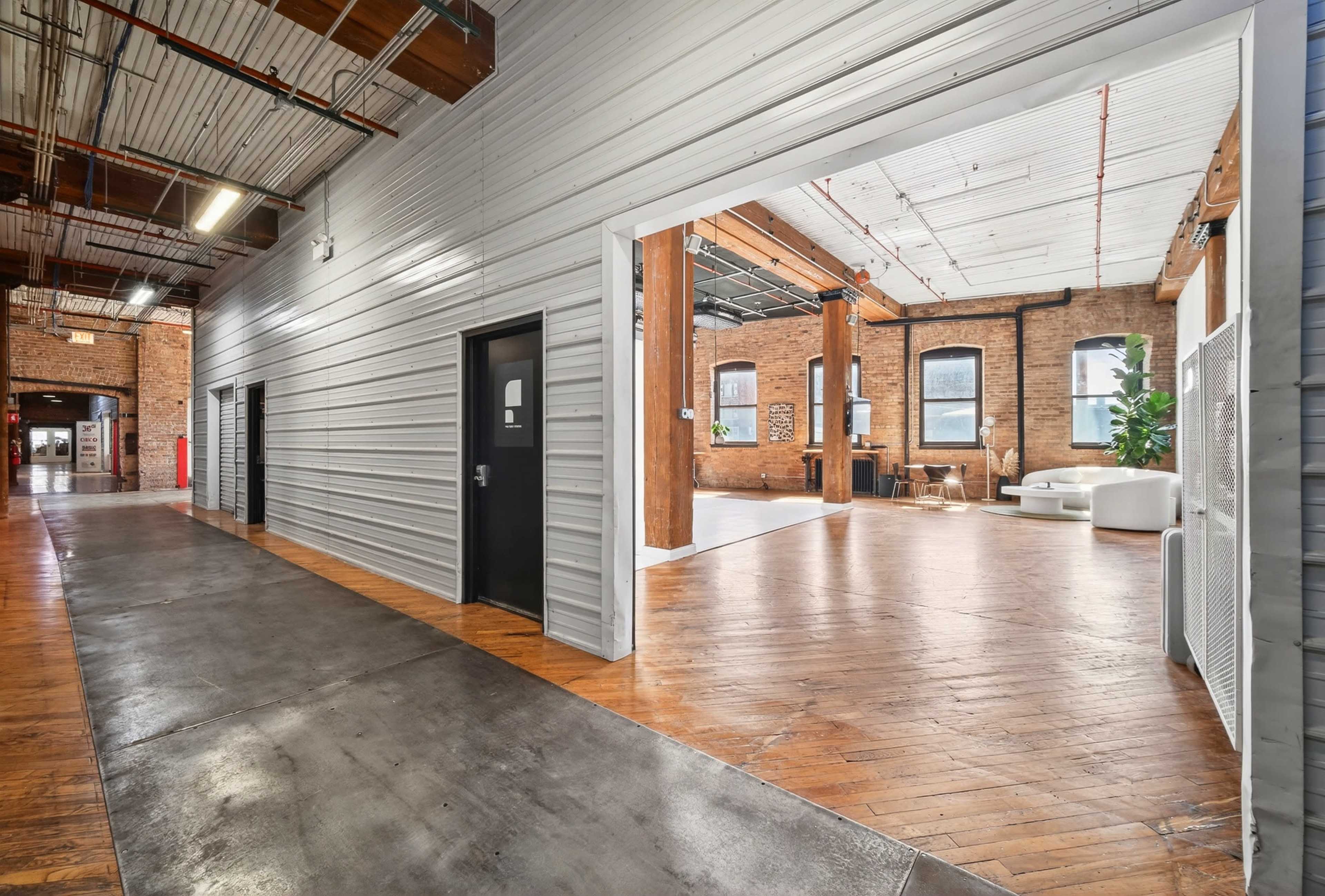 The image shows a long corridor with a polished concrete floor leading to an open space featuring wooden flooring and large windows.