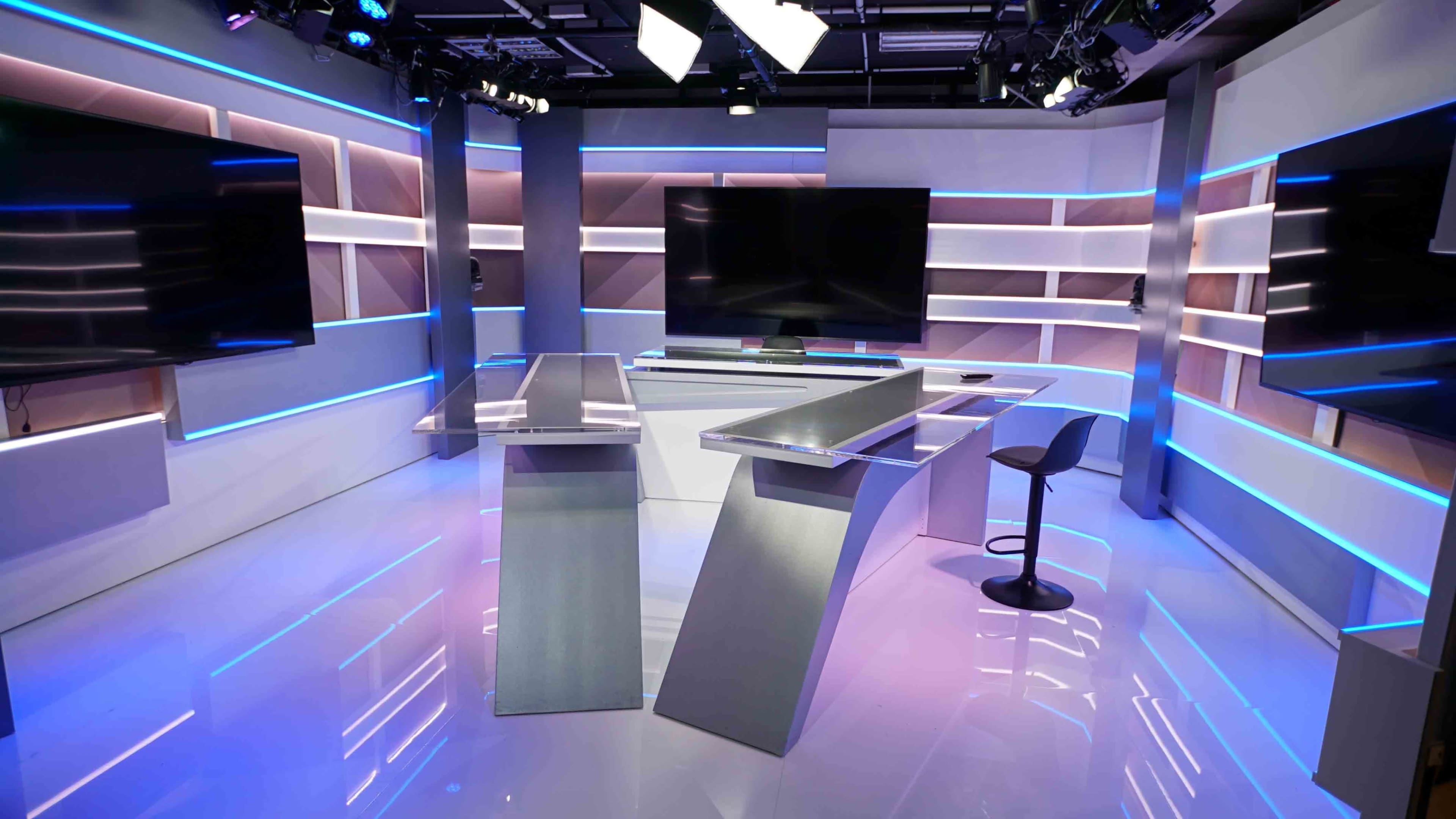 The image shows a modern television news studio with sleek, minimalist furniture and illuminated shelving.