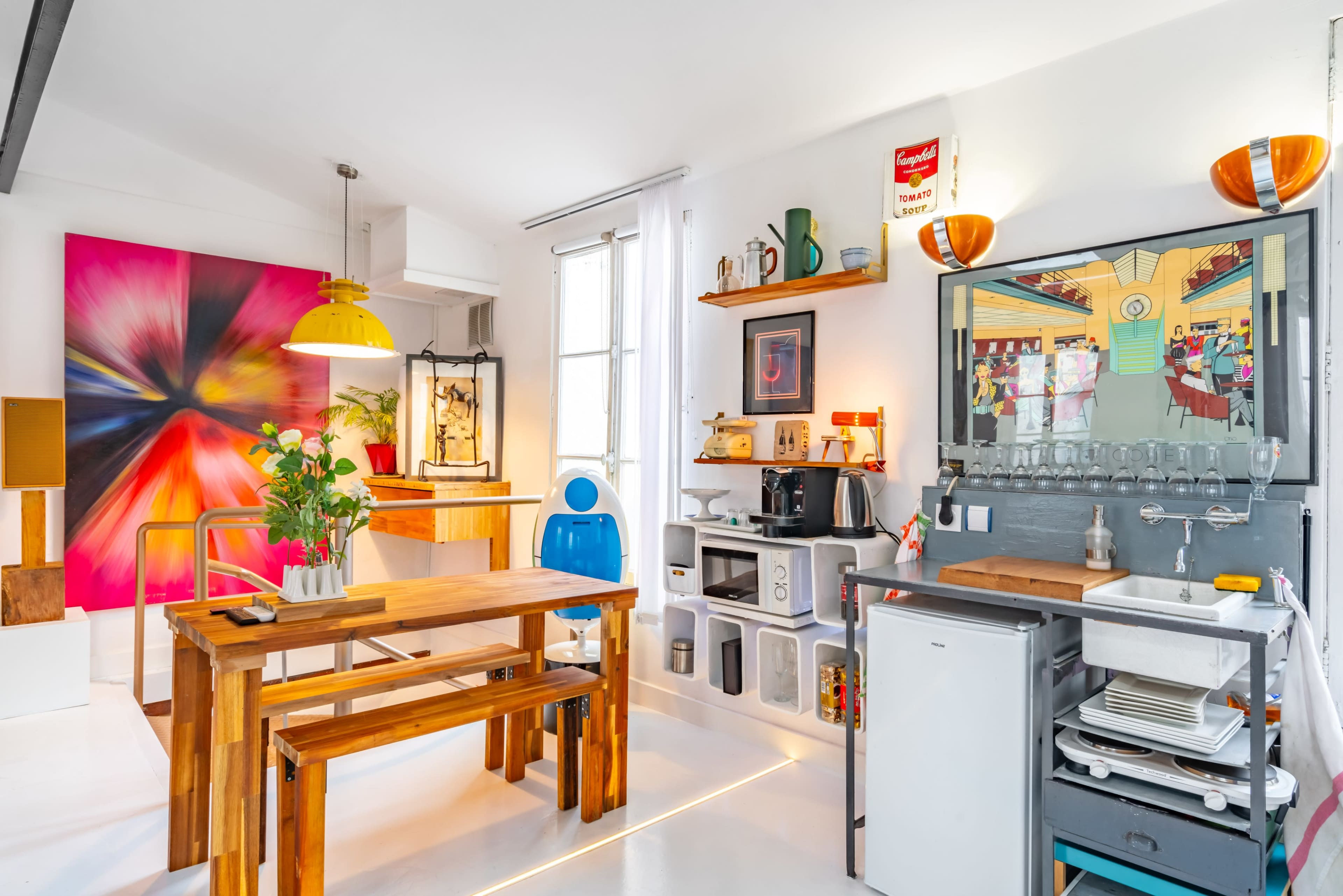 A bright, modern kitchen setup featuring a wooden dining table and chairs, colorful wall art, and a compact kitchen area with a sink and storage shelves.