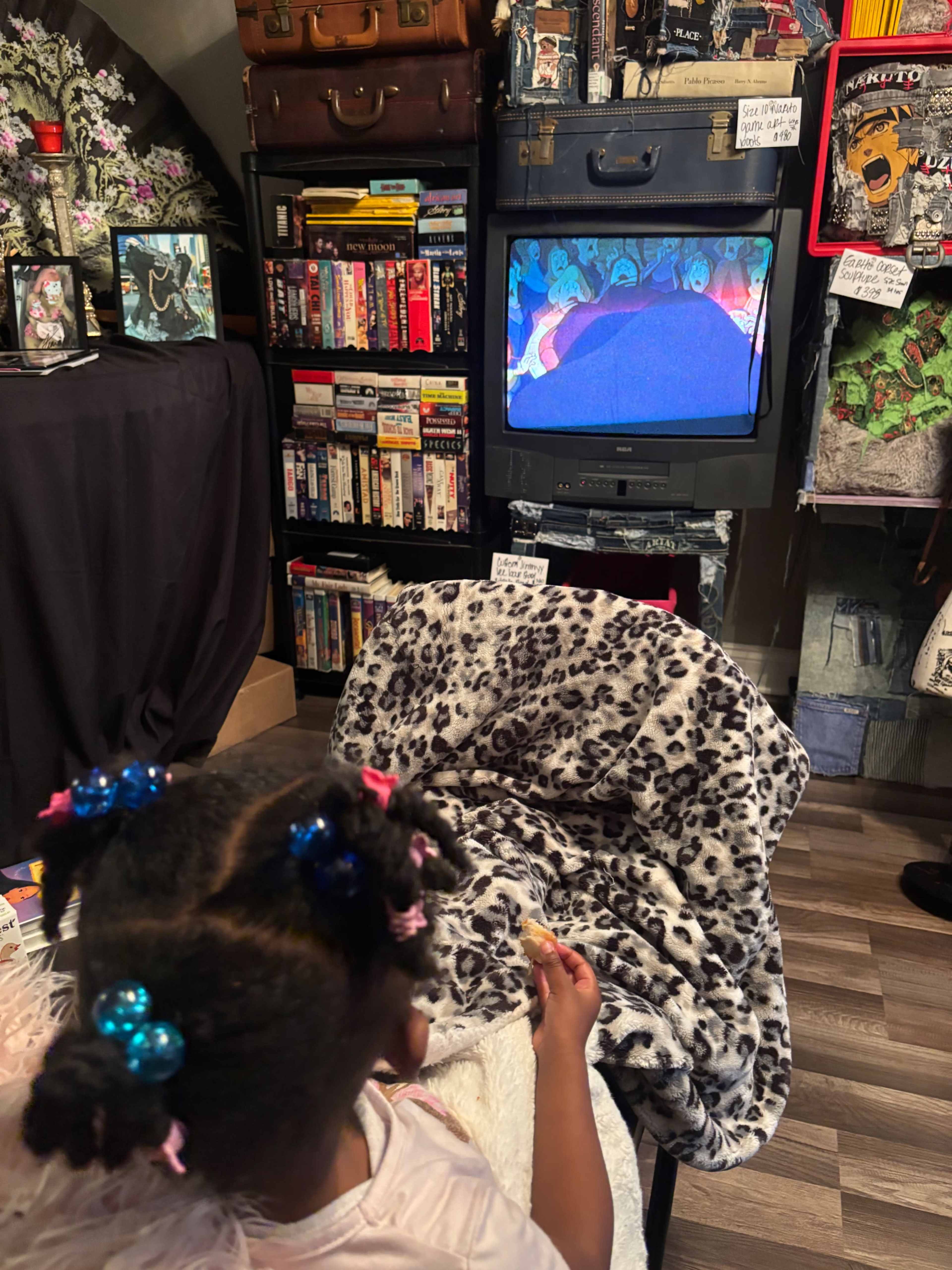 A child sits on a chair covered with a leopard-print blanket, watching an animated show on an old television surrounded by shelves filled with various items and media.