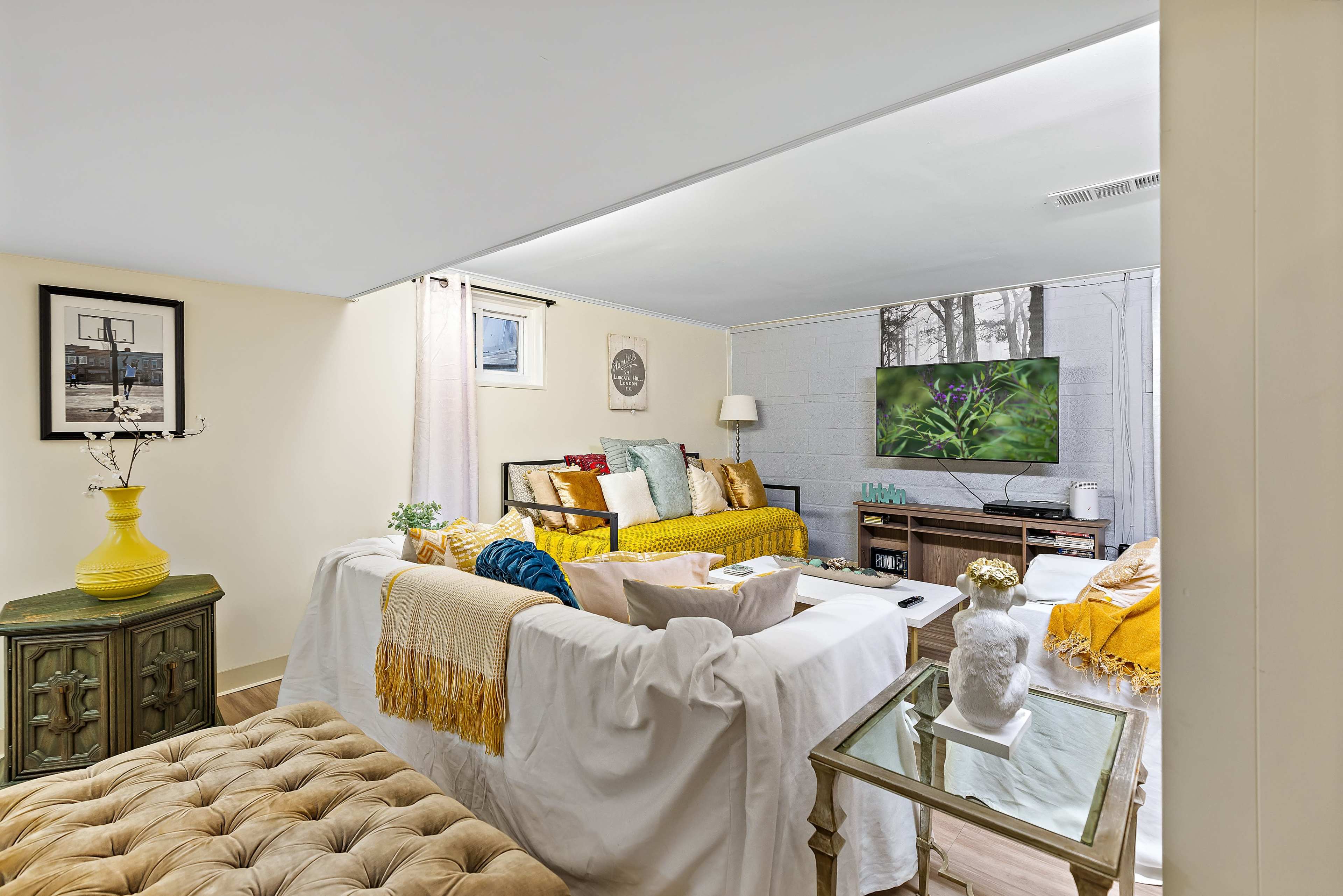 A cozy basement living area features a sectional sofa, a large TV, and various decorative elements, including colorful throw pillows and a small table.
