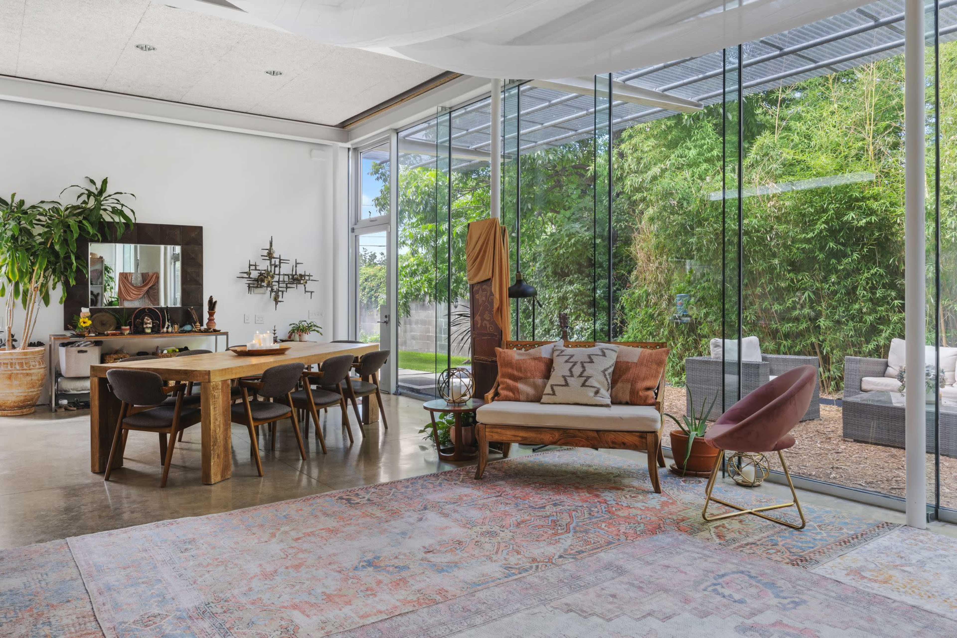 A modern dining area features a wooden table with black chairs, a sofa with decorative pillows, and large glass windows overlooking a green outdoor space.
