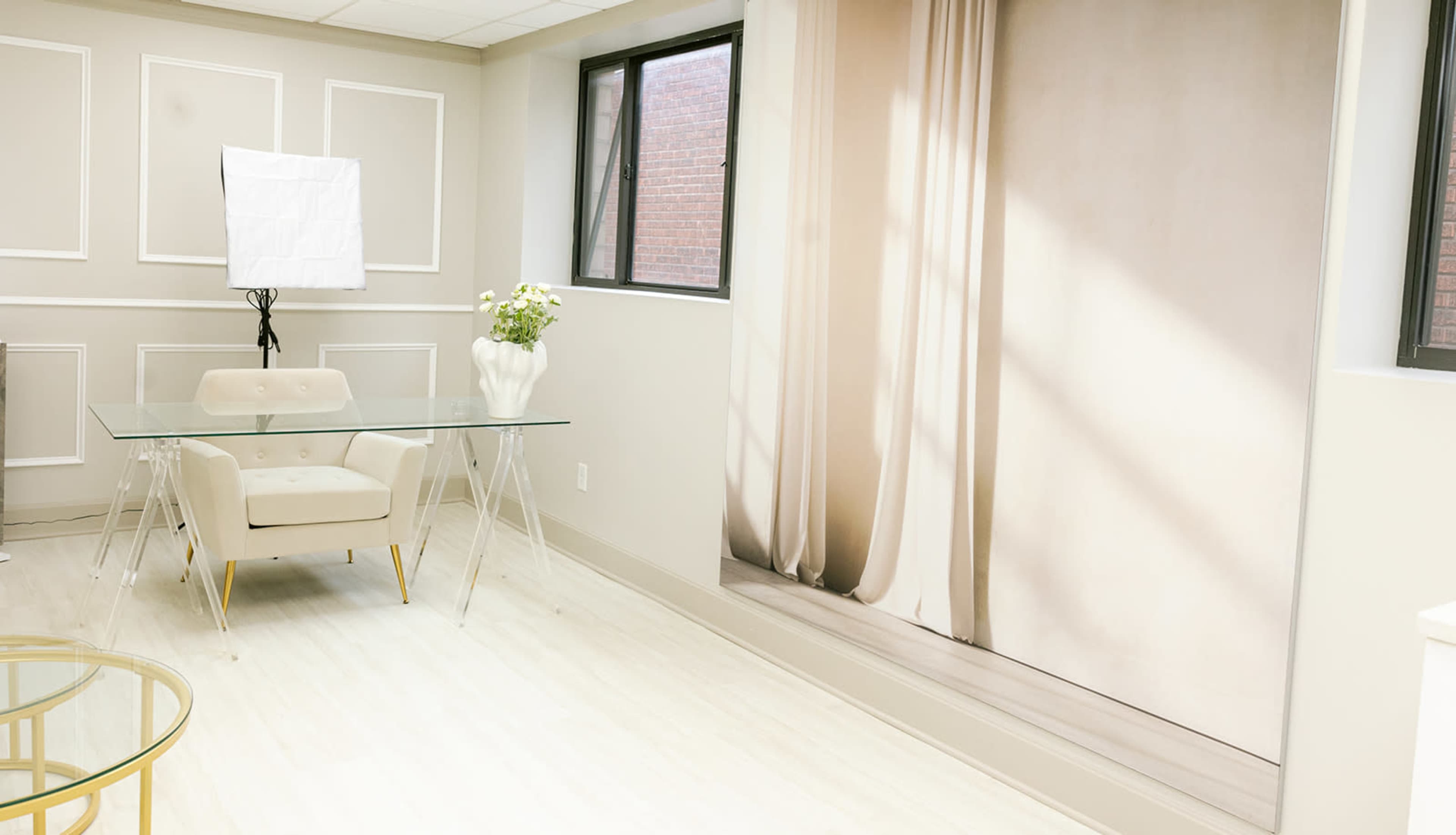 The image depicts a modern office space featuring a glass table and a light-colored chair, with a large curtain backdrop on one wall.