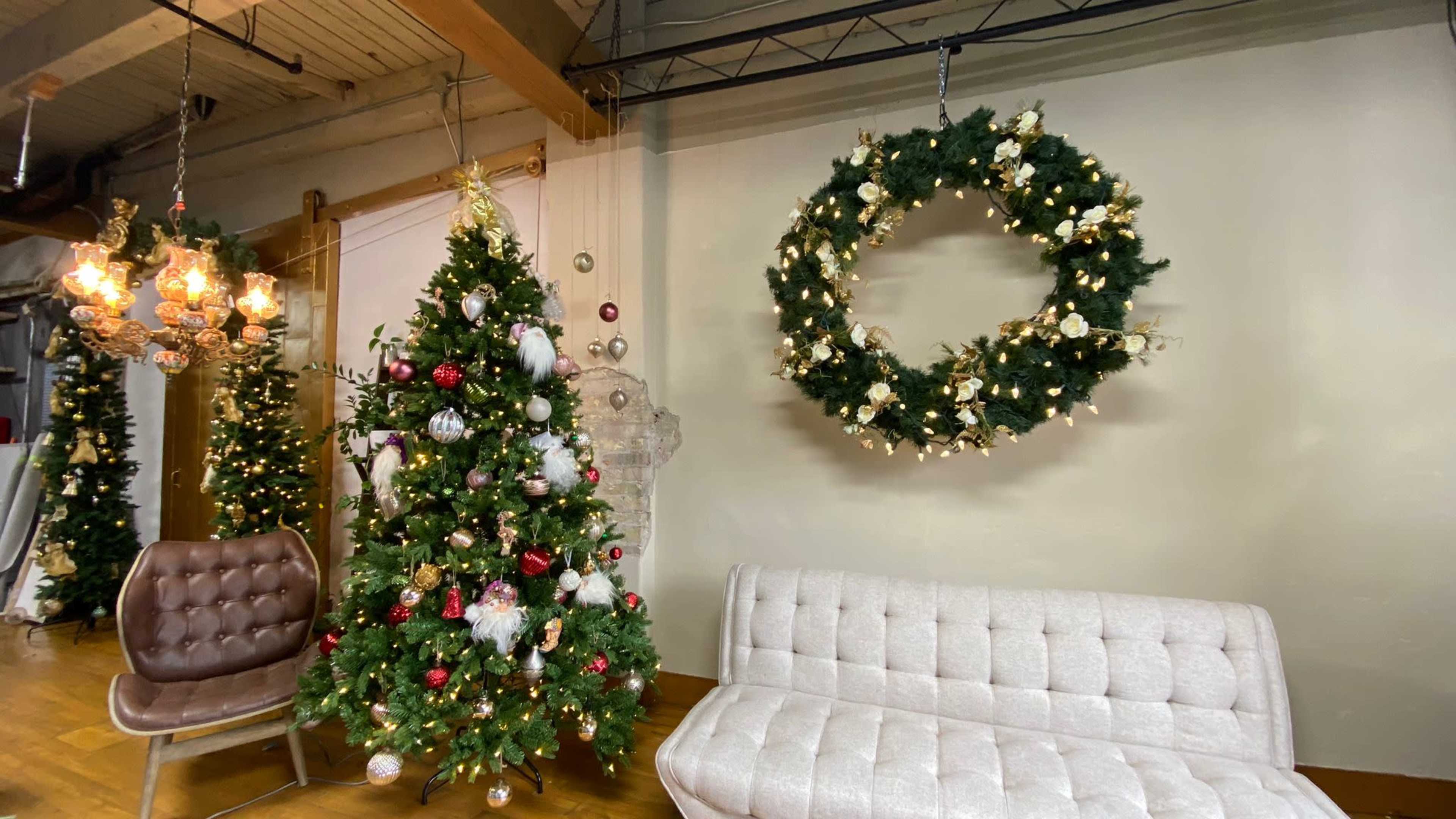 A decorated Christmas tree stands beside a modern white sofa in a rustic indoor setting with a large wreath hanging on the wall.