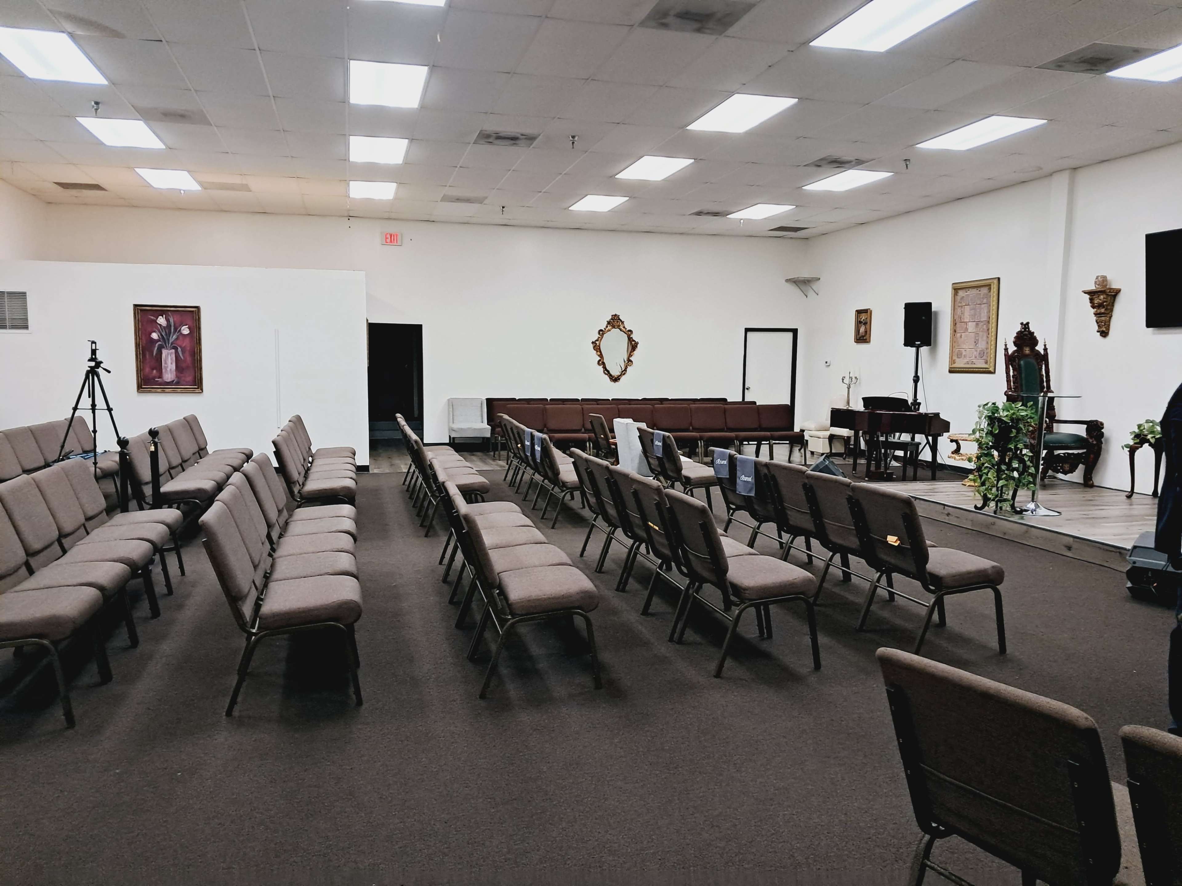 The image shows an indoor space set up with rows of metal-framed chairs arranged for an event, with a stage area visible at one end.