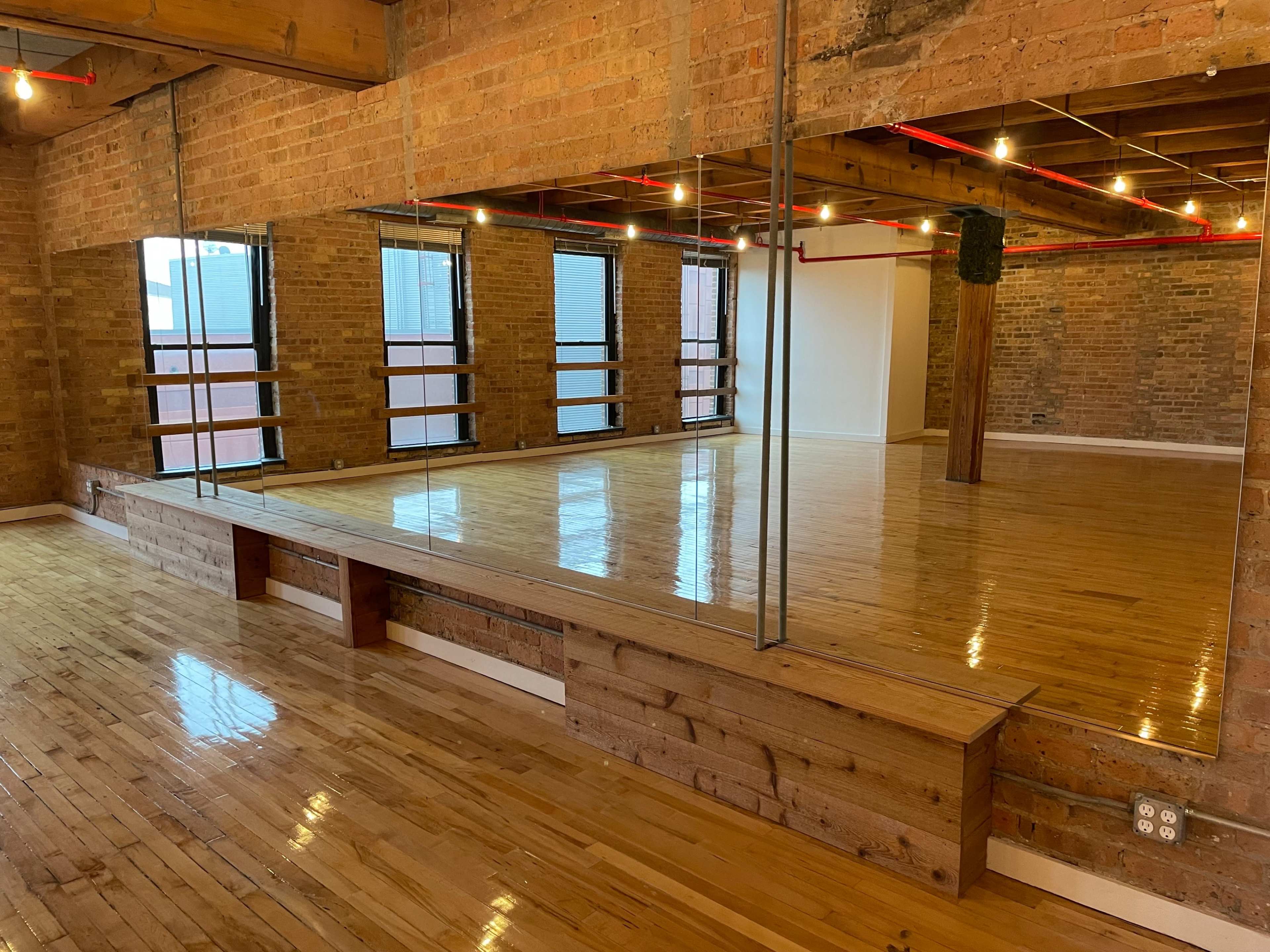 The image shows a spacious dance studio with polished wooden floors, exposed brick walls, and large mirrors reflecting the room's interior.