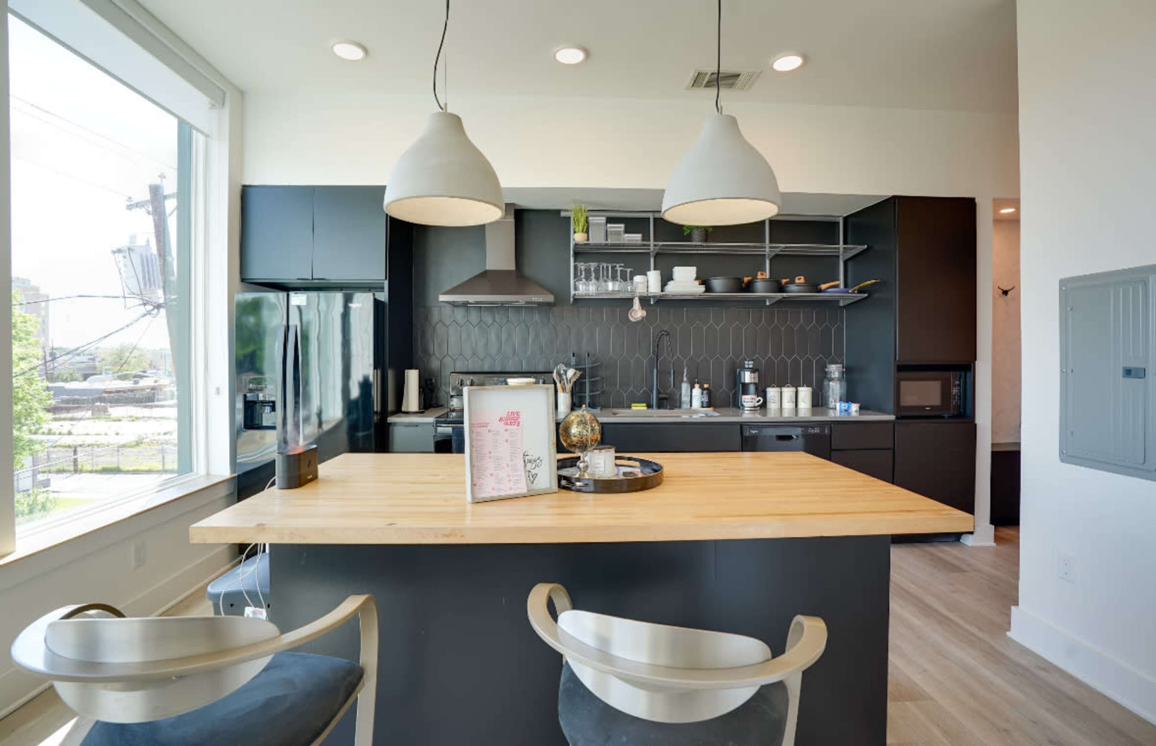 A modern kitchen features a central wooden island with two chairs, black cabinetry, shelves, and pendant lighting.