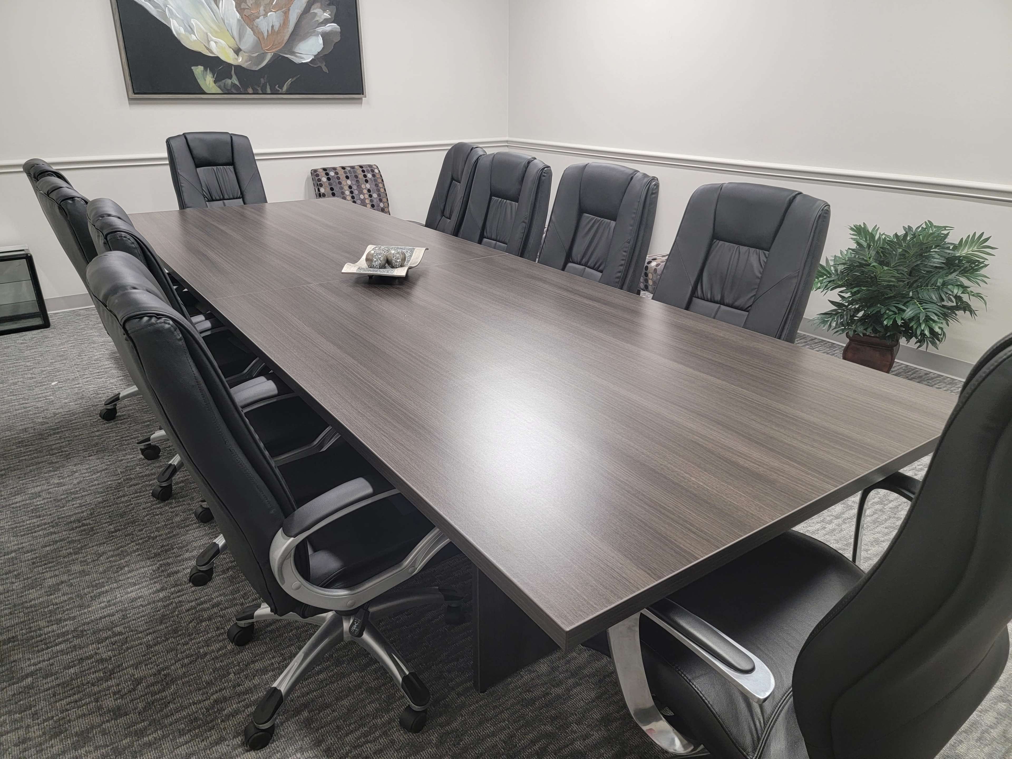 A long conference table with black leather chairs surrounds it, and a small decorative item sits in the center, with a plant in the corner and a framed floral artwork on the wall.