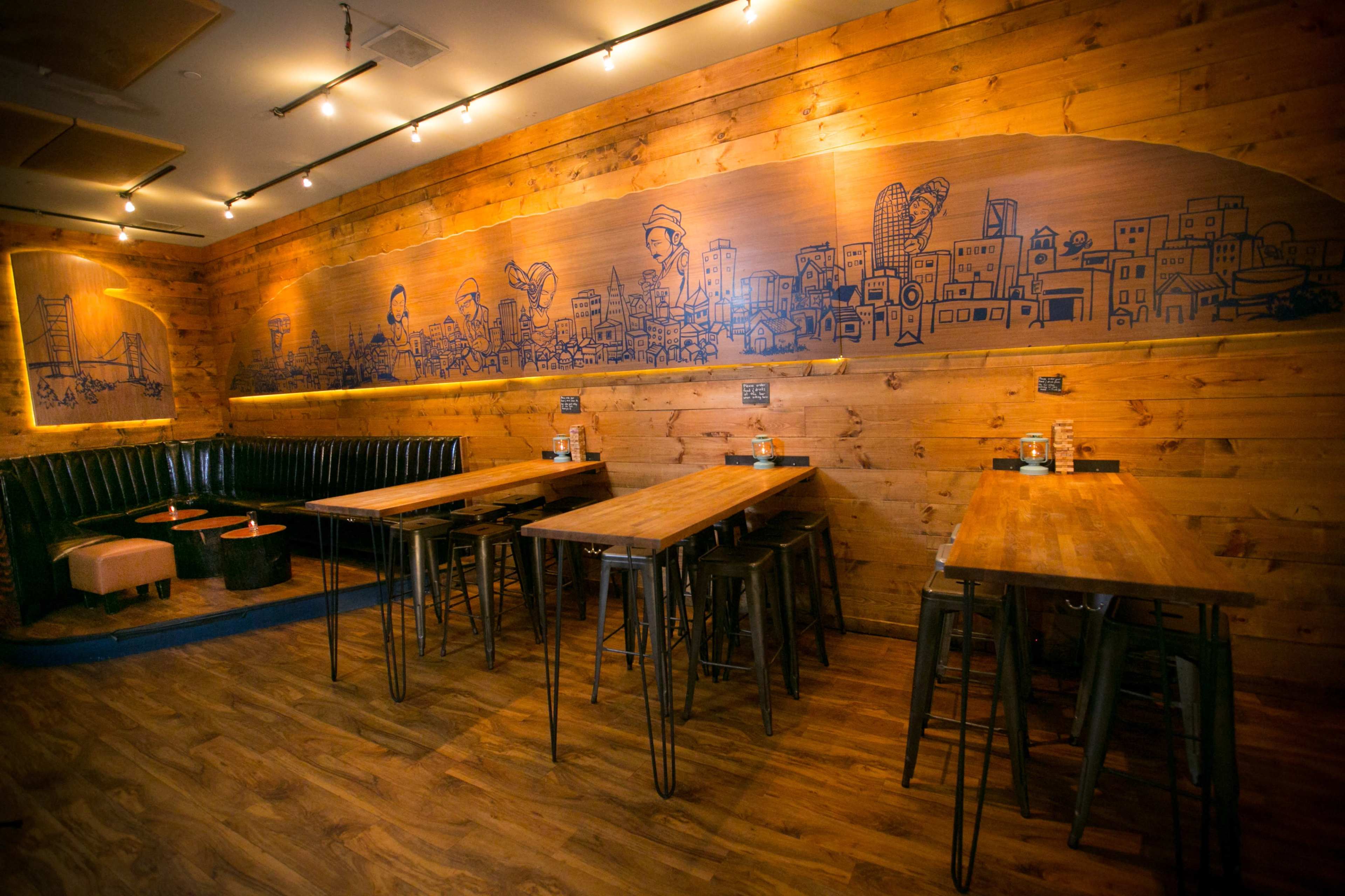 A wooden-themed restaurant interior with multiple wooden tables, tall stools, and a mural depicting a cityscape on the wall.