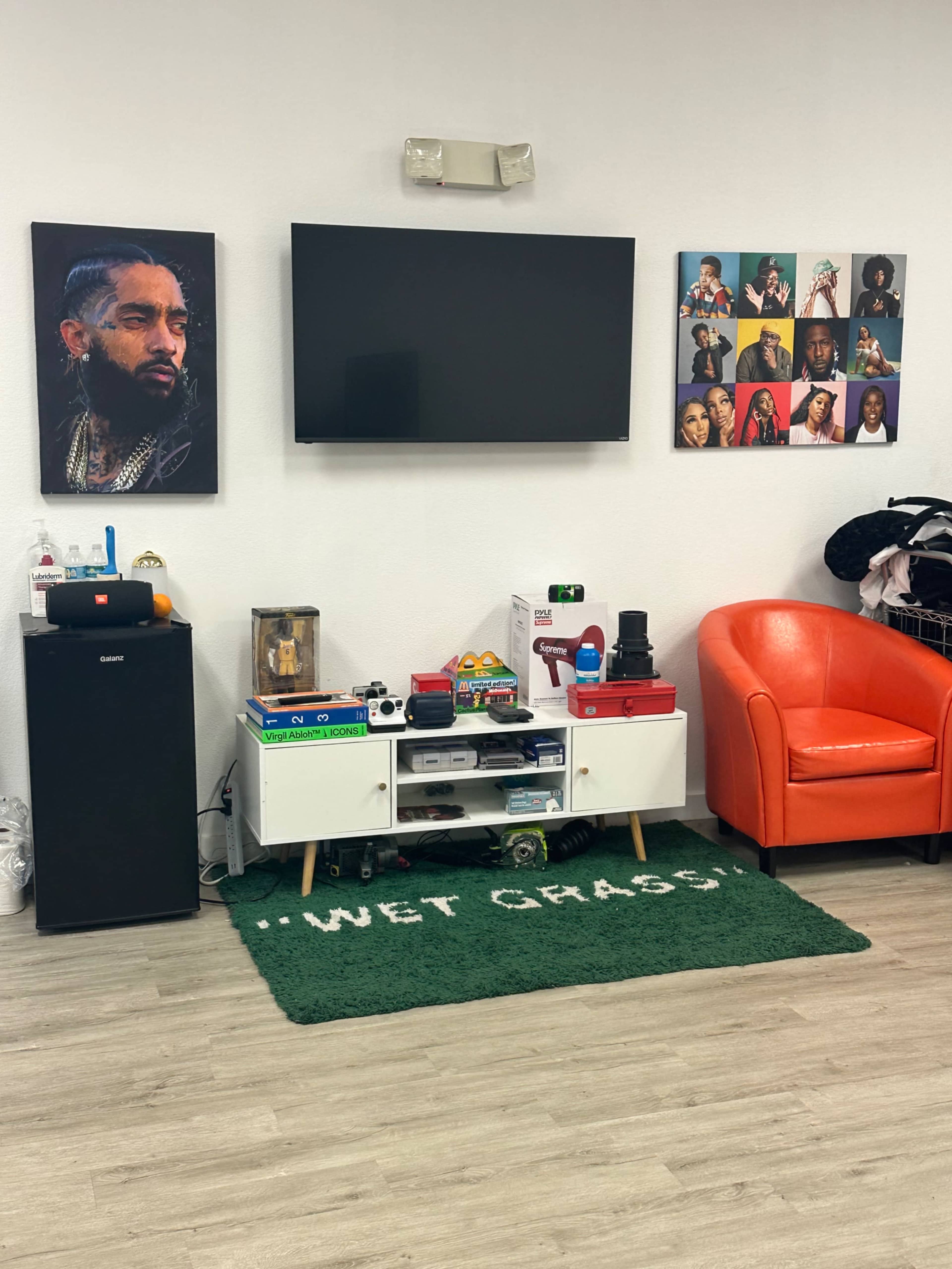 A room with a large flat-screen TV mounted on the wall, a black mini-fridge, a white entertainment unit filled with various items, an orange chair, and several framed pictures of musicians displayed on the wall.
