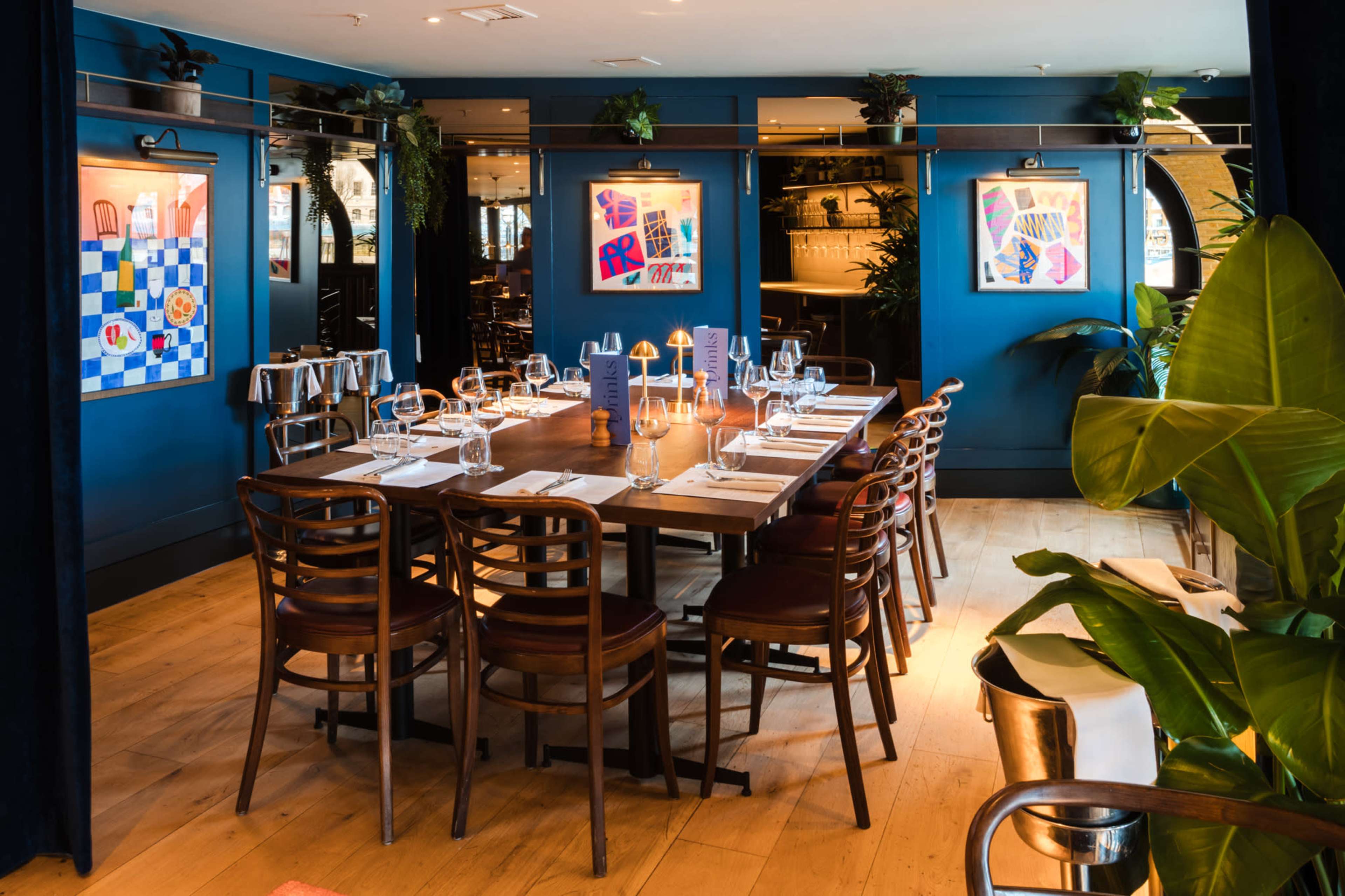 The image shows a dining room with a long wooden table set for a meal, surrounded by wooden chairs and adorned with colorful artwork on the blue walls.