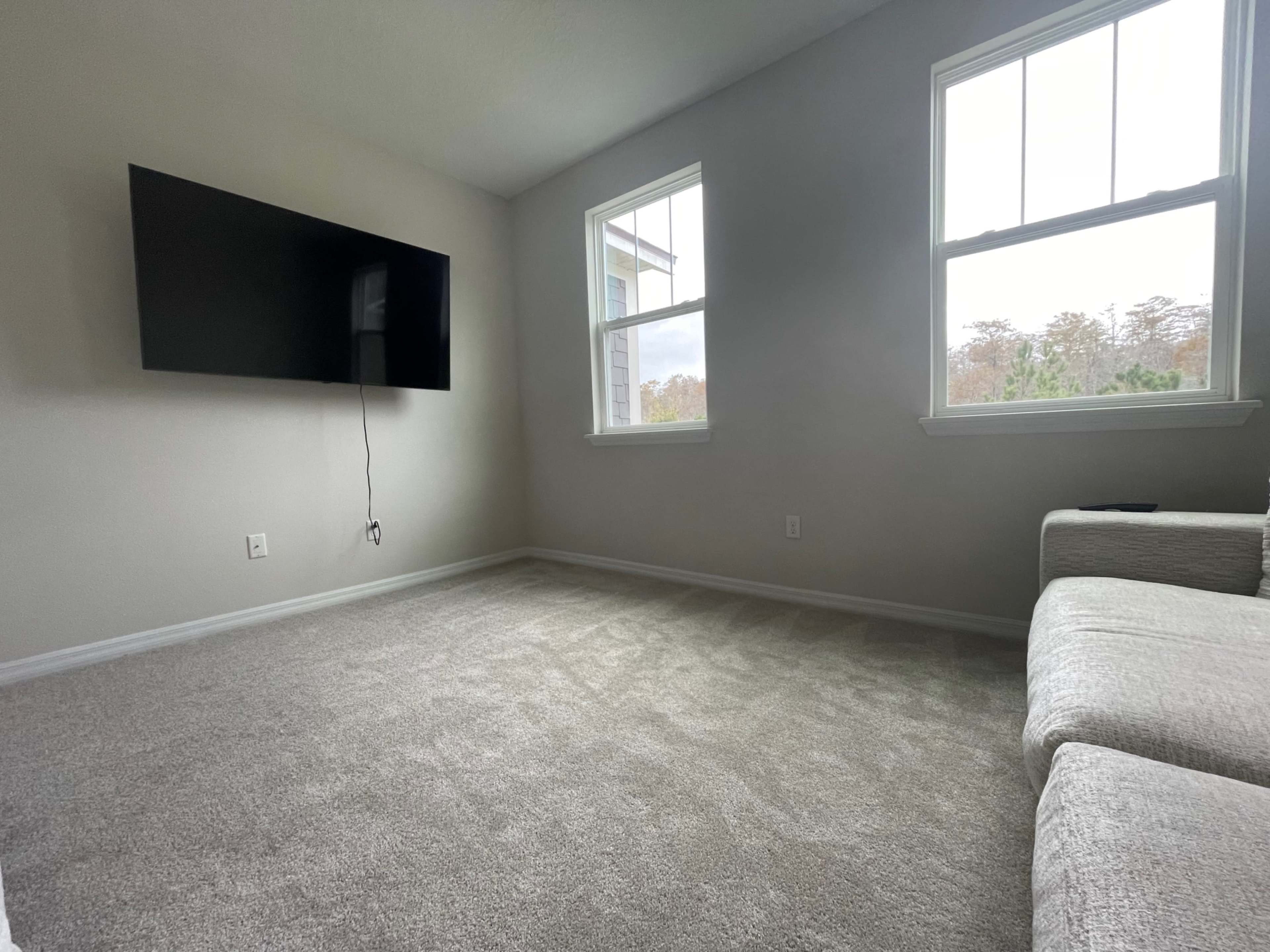 A sparsely furnished living room with gray carpet, a wall-mounted television, two windows, and a light-colored sofa.