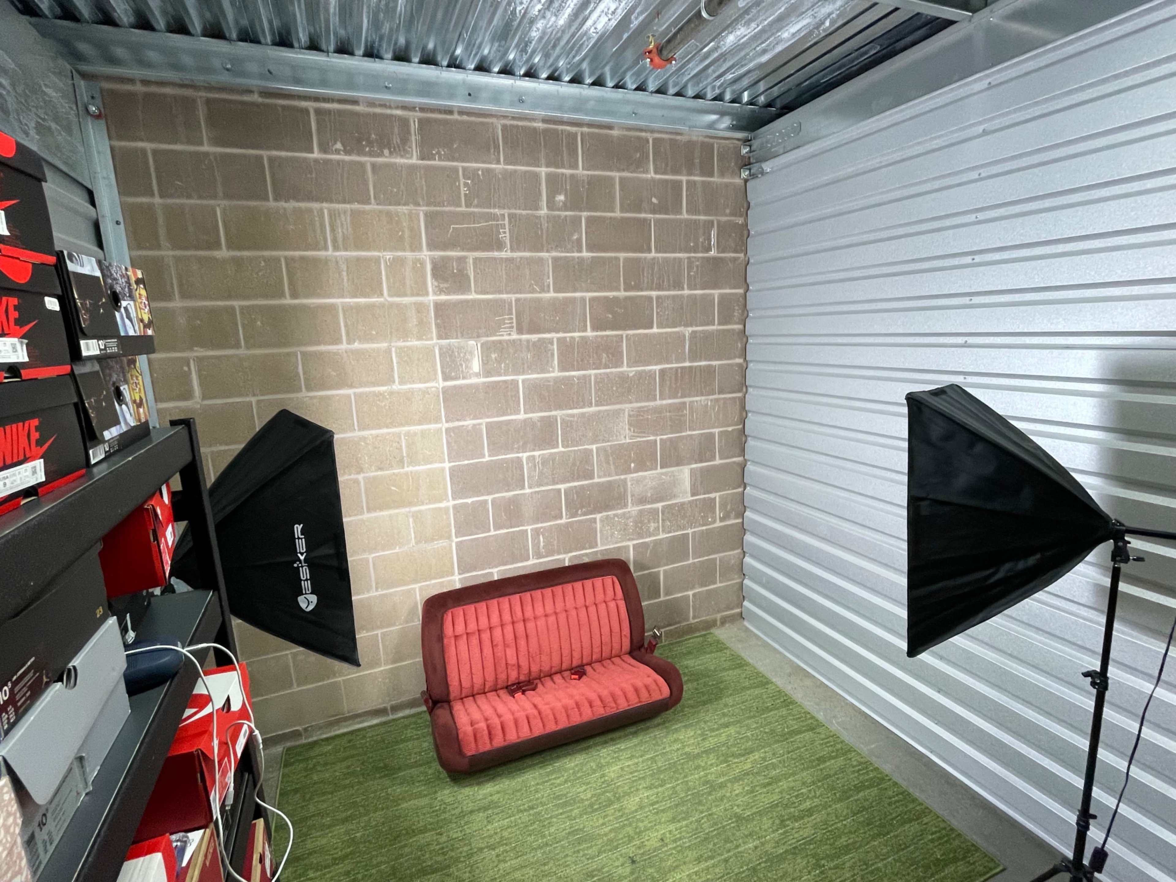 The image shows a small storage unit with a red couch, two softbox lights, and organized shoe boxes stacked on shelves against the wall.