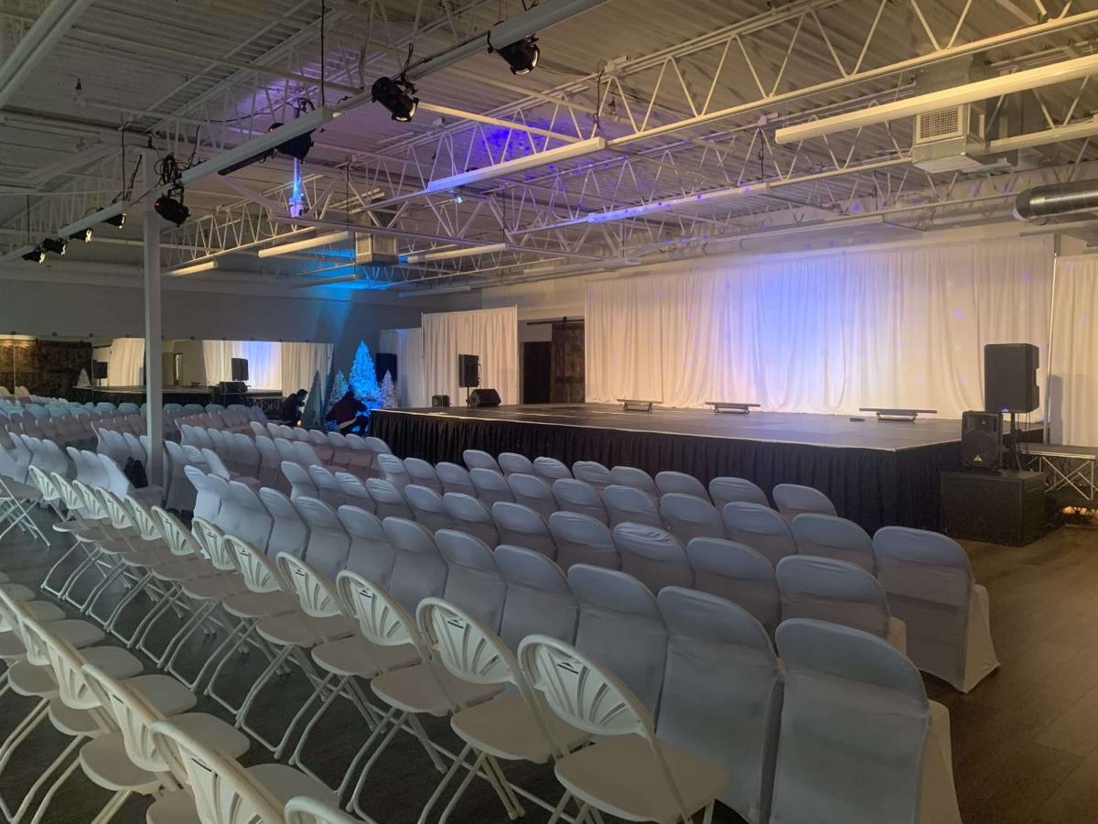 A spacious event hall set up with rows of white chairs facing a stage adorned with a white backdrop and dim lighting.