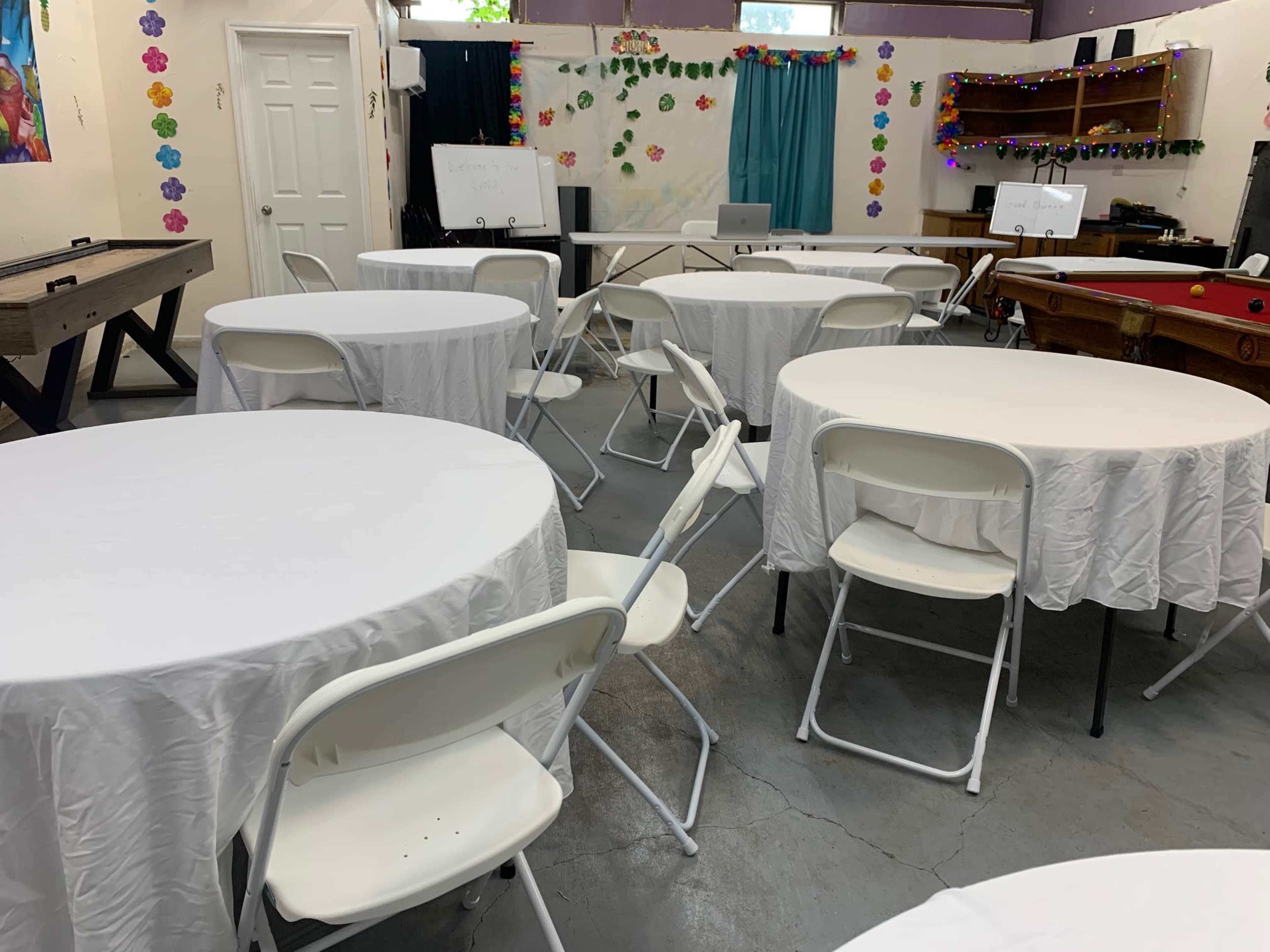 The image shows a room set up for an event, featuring several round tables covered with white tablecloths and surrounded by white folding chairs.