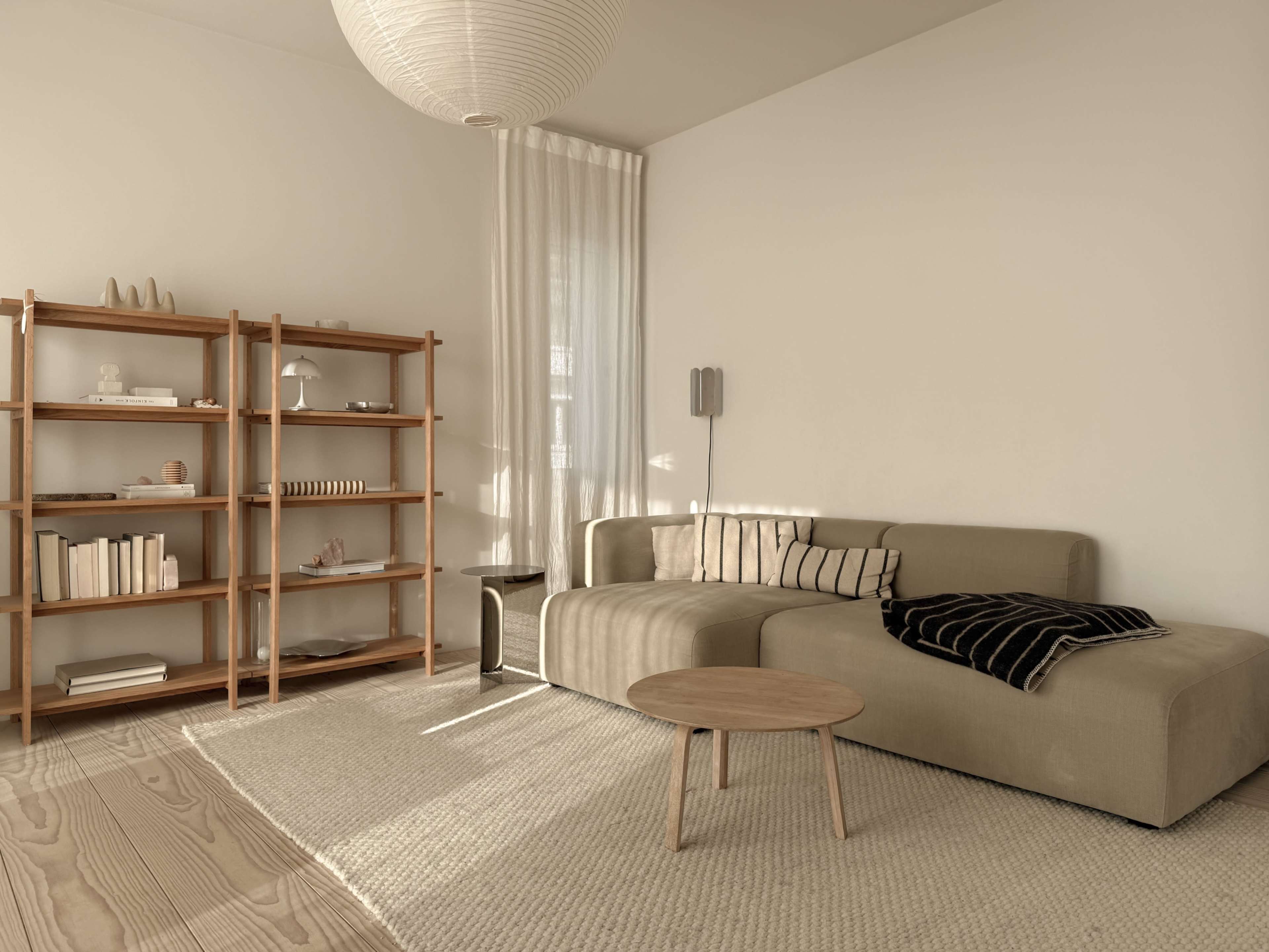 A minimalist living room features a light-colored sofa, a wooden bookshelf filled with decor, and a round coffee table on a textured rug.