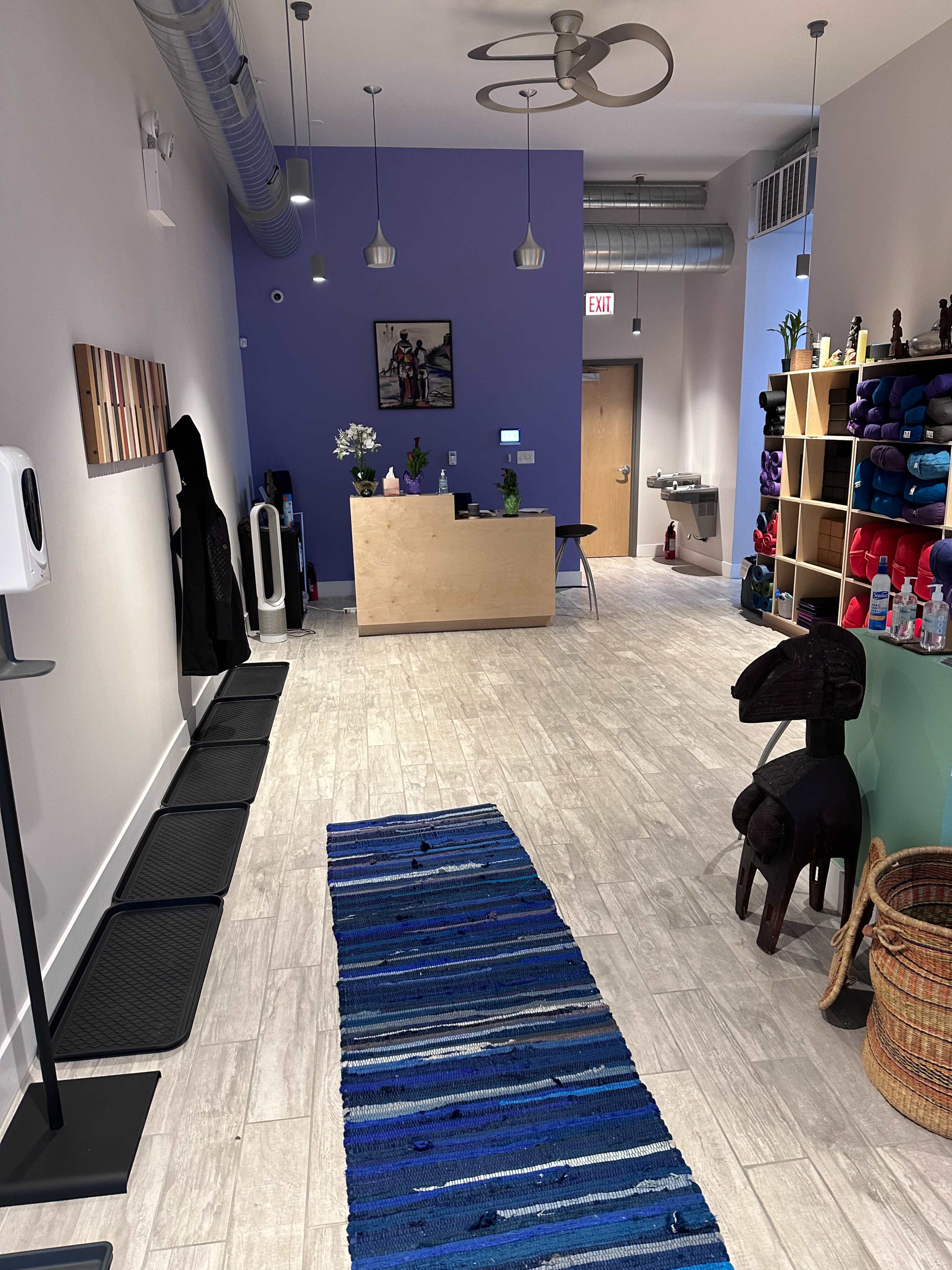 The image depicts a brightly lit reception area with a wooden counter, shelves displaying colorful items, and a striped blue rug leading towards the entrance.