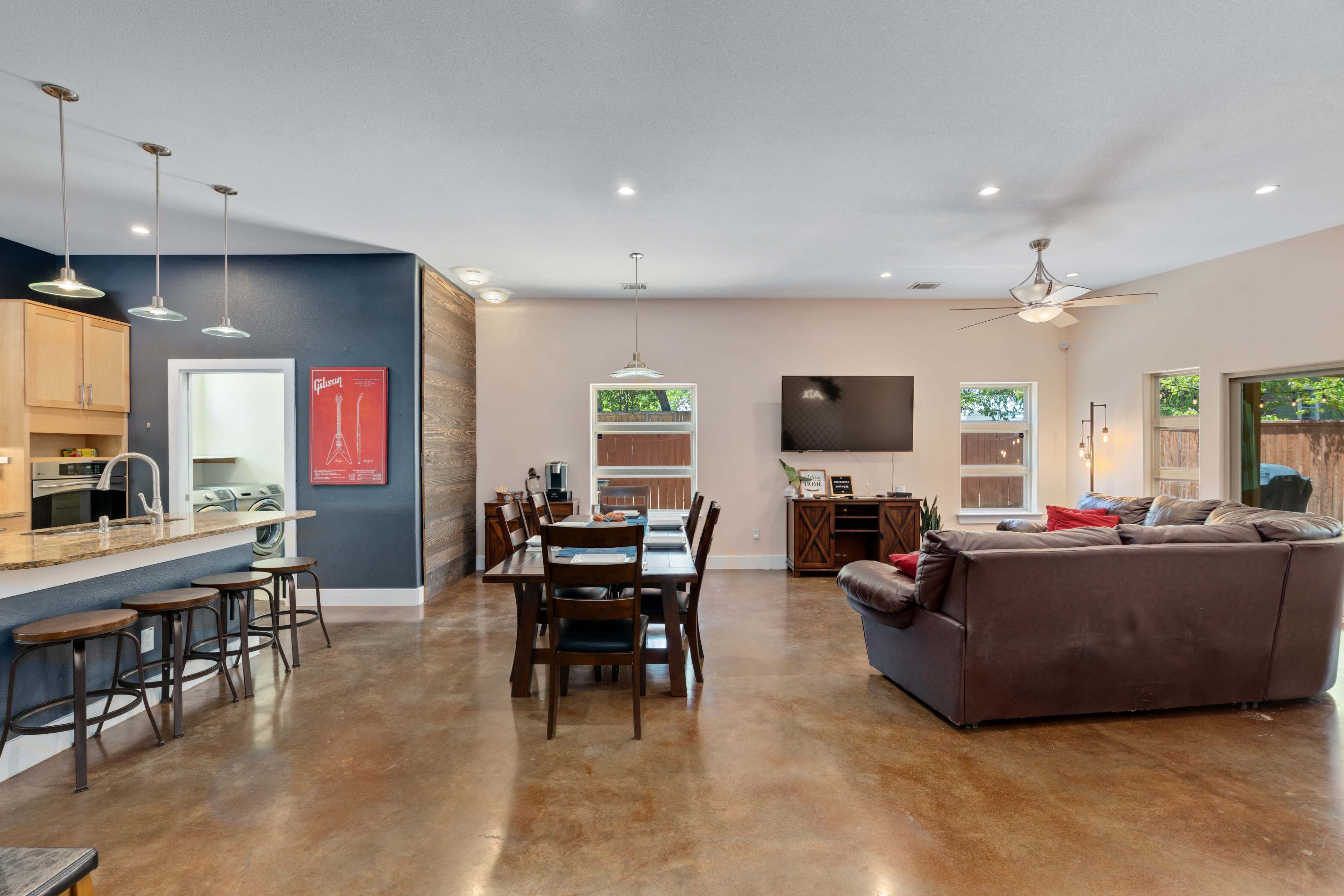 A spacious living and dining area featuring a wooden dining table, a dark sofa, and a kitchen bar with stools.