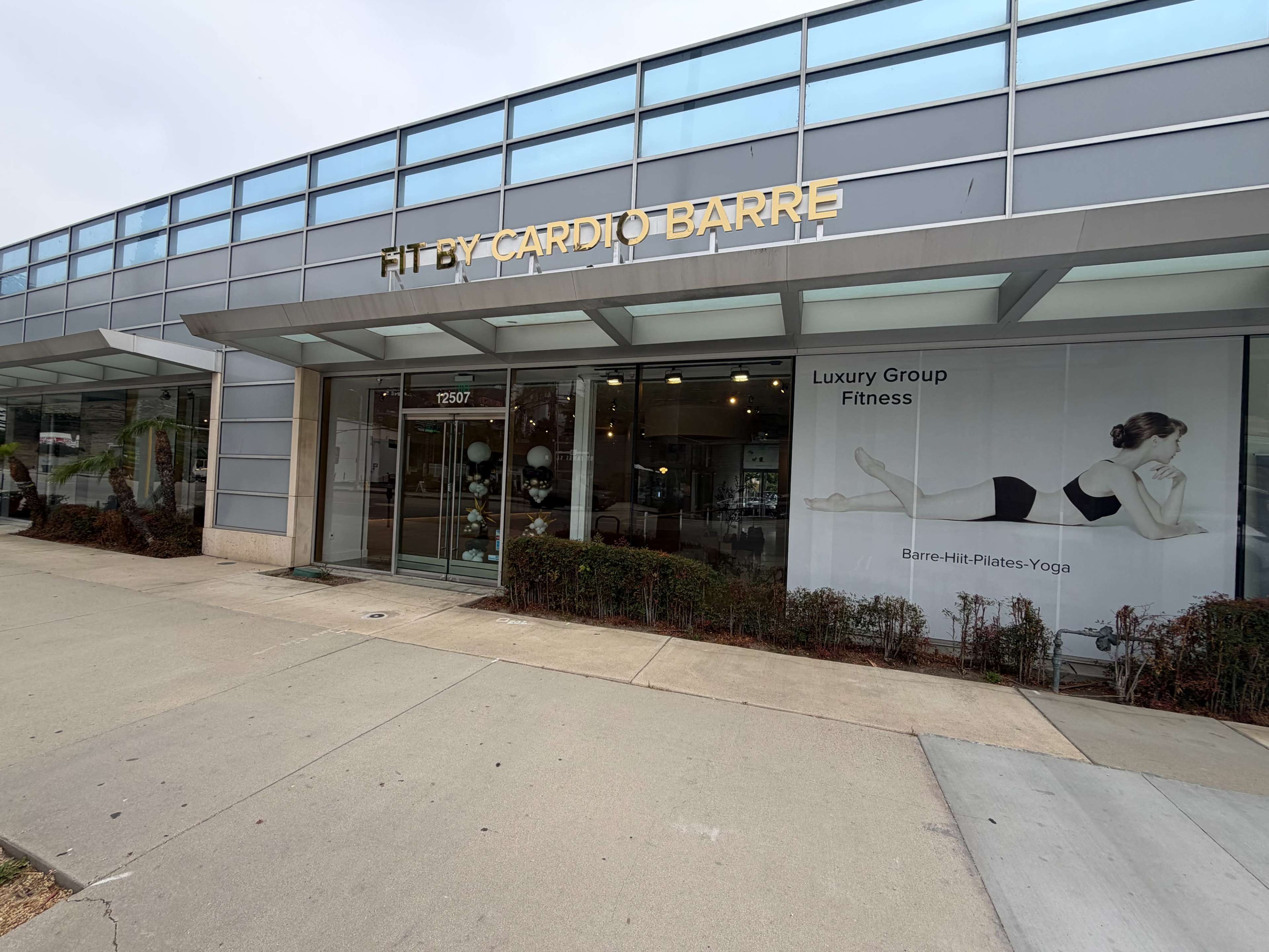The image shows a storefront for "Fit by Cardio Barre," featuring large glass windows and a promotional banner highlighting "Luxury Group Fitness" with a depiction of a woman in a workout pose.