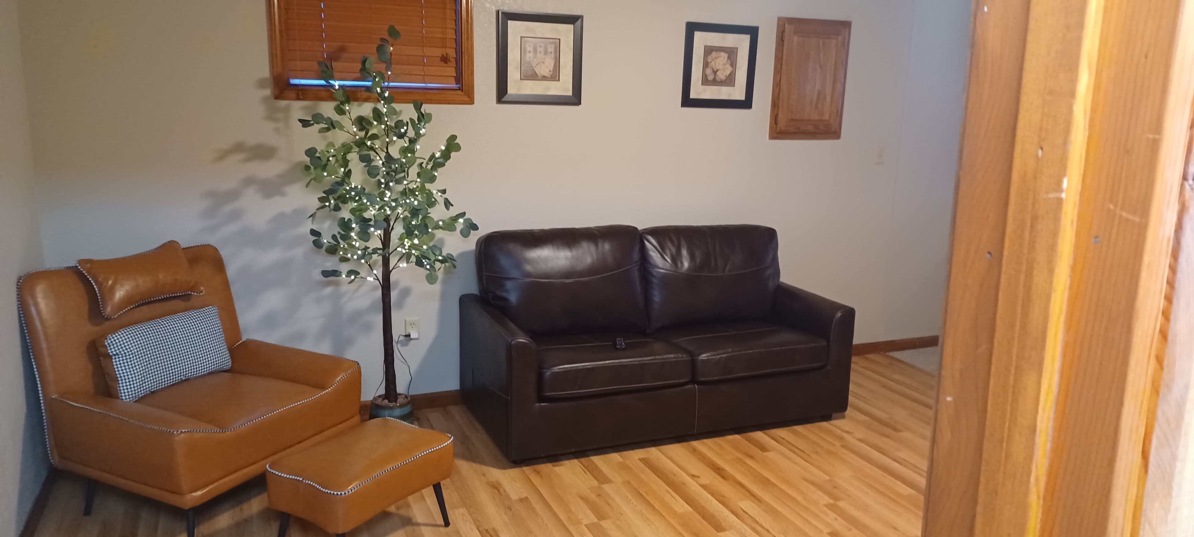 The image shows a living room with a brown leather sofa, a tan armchair with an ottoman, and a decorative plant next to framed artwork on the wall.