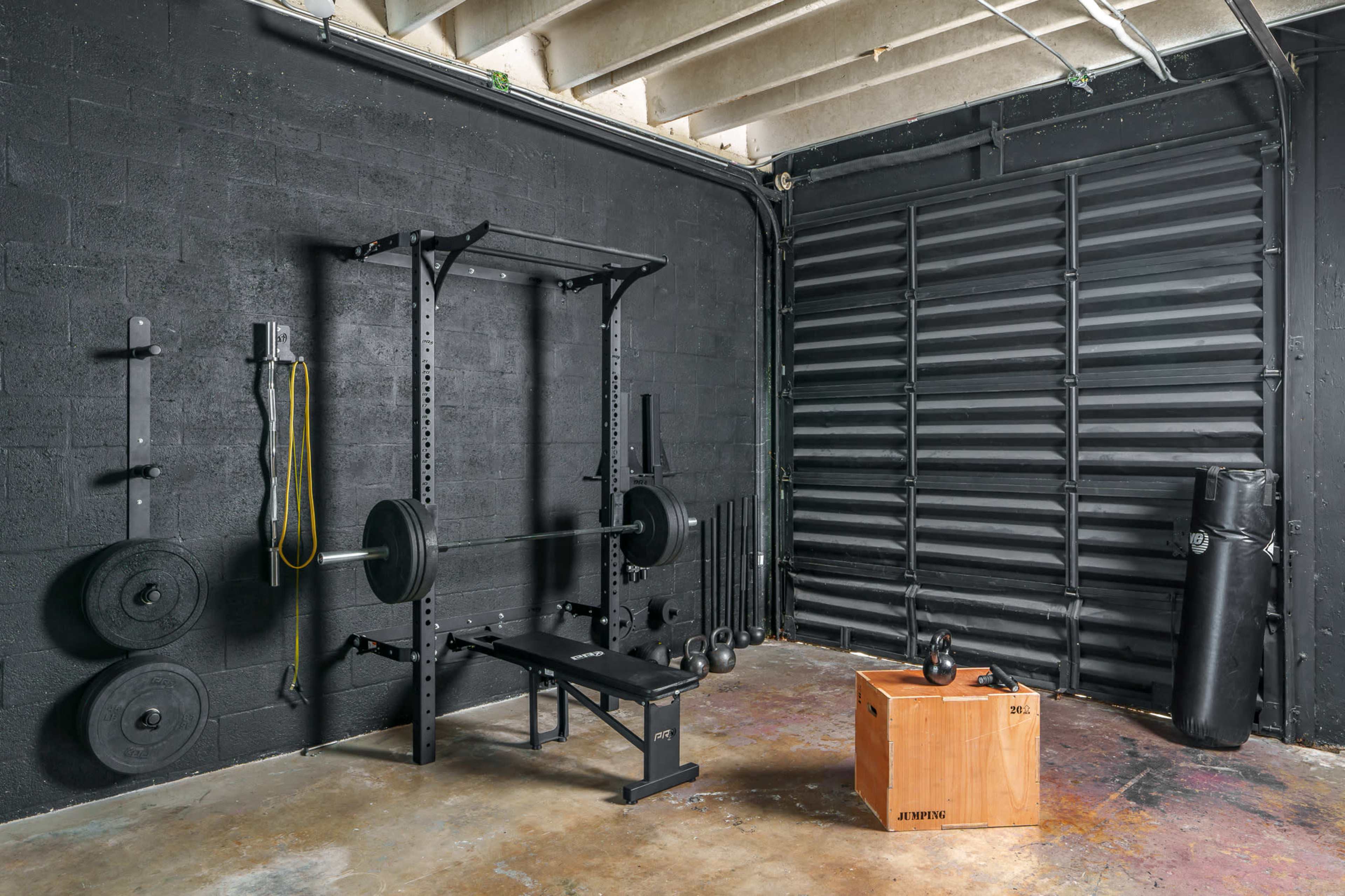 The image shows a gym setup in a black-walled room featuring a weightlifting rack with a barbell, a bench, a wooden plyometric box, a kettlebell, and a punching bag.