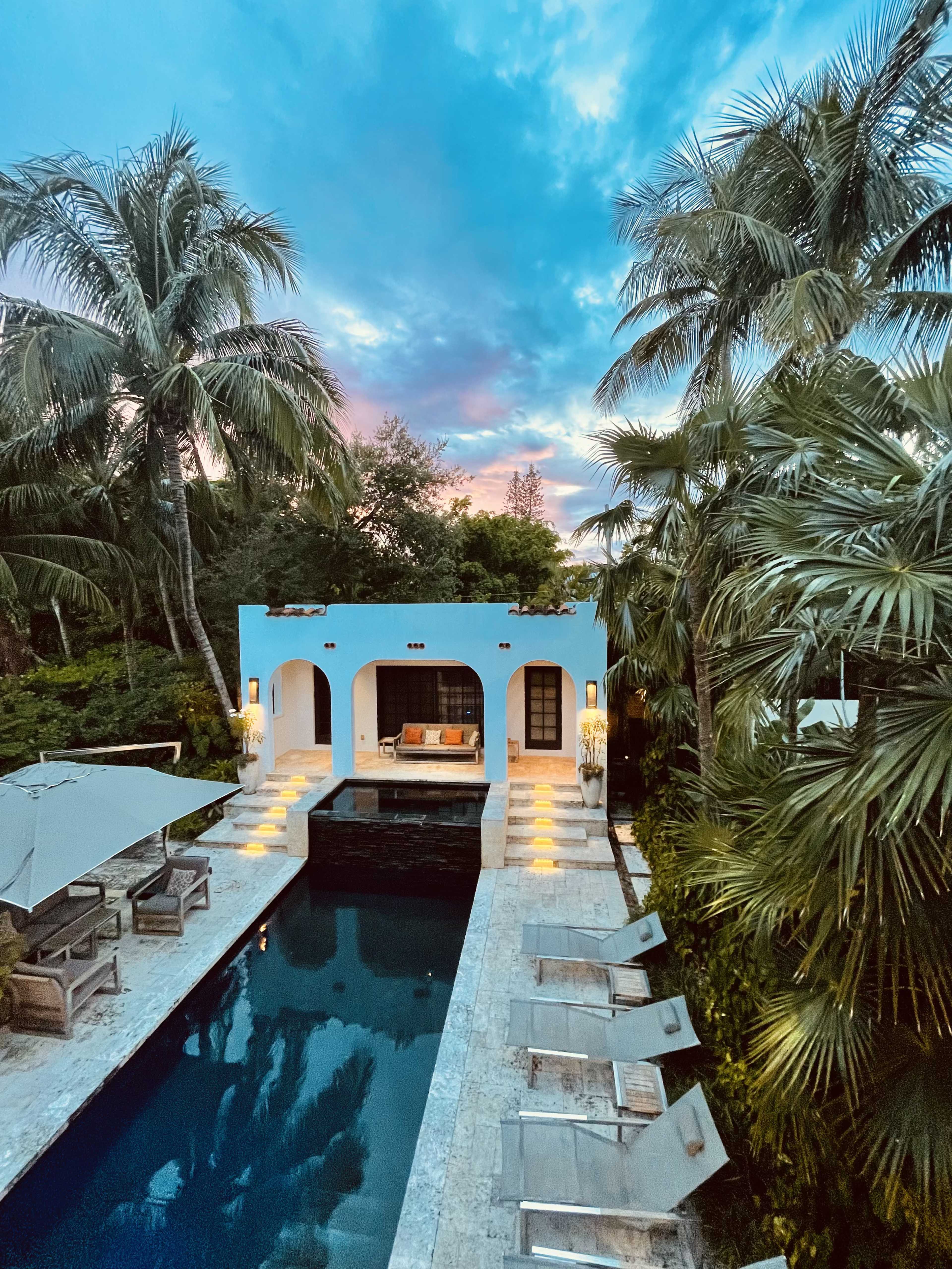 A pool surrounded by palm trees, with a blue building and lounge chairs nearby, illuminated by pathway lights at dusk.