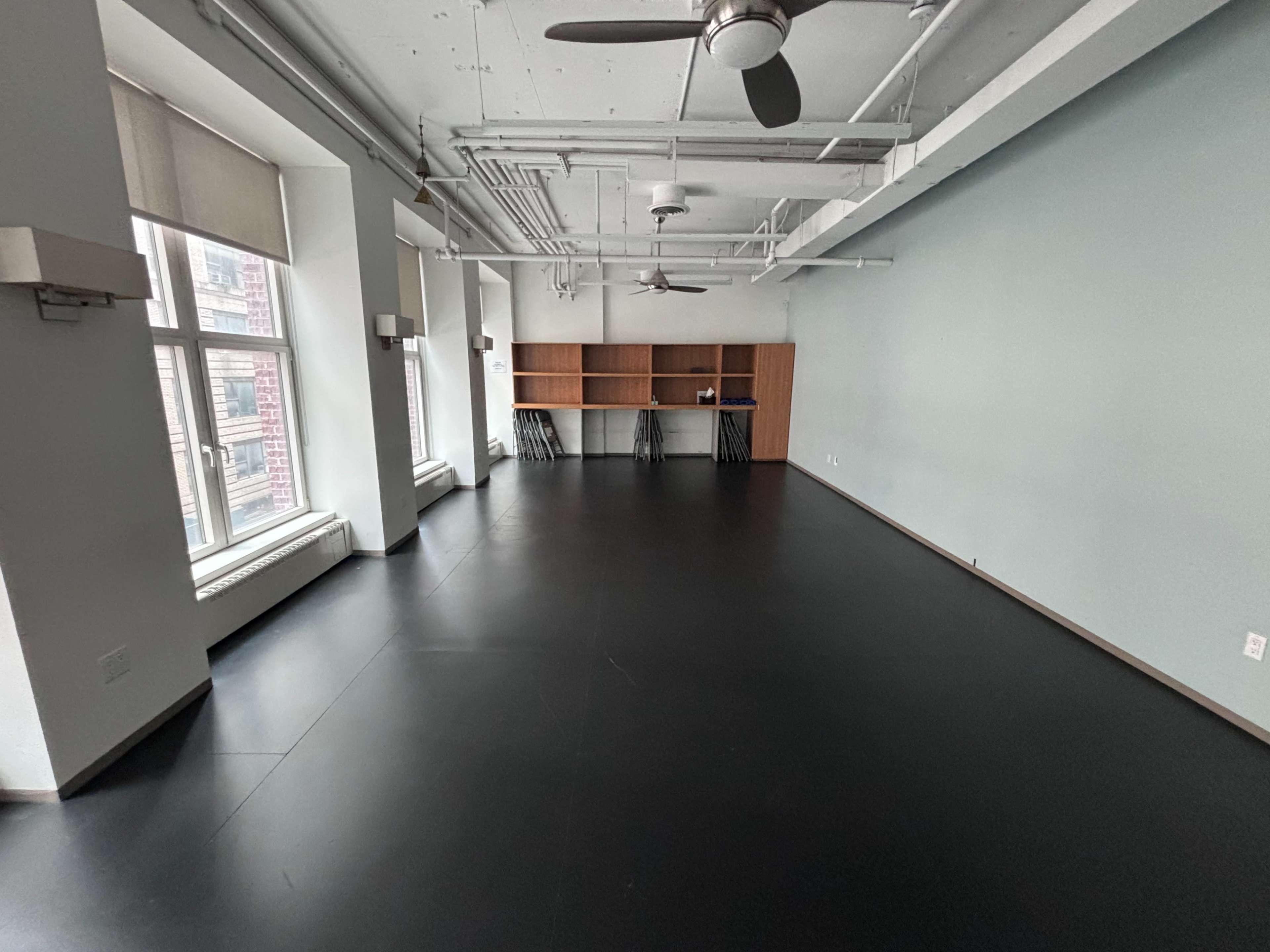 The image shows a spacious, empty room with a smooth black floor, two large windows, and a wooden shelving unit against the wall.