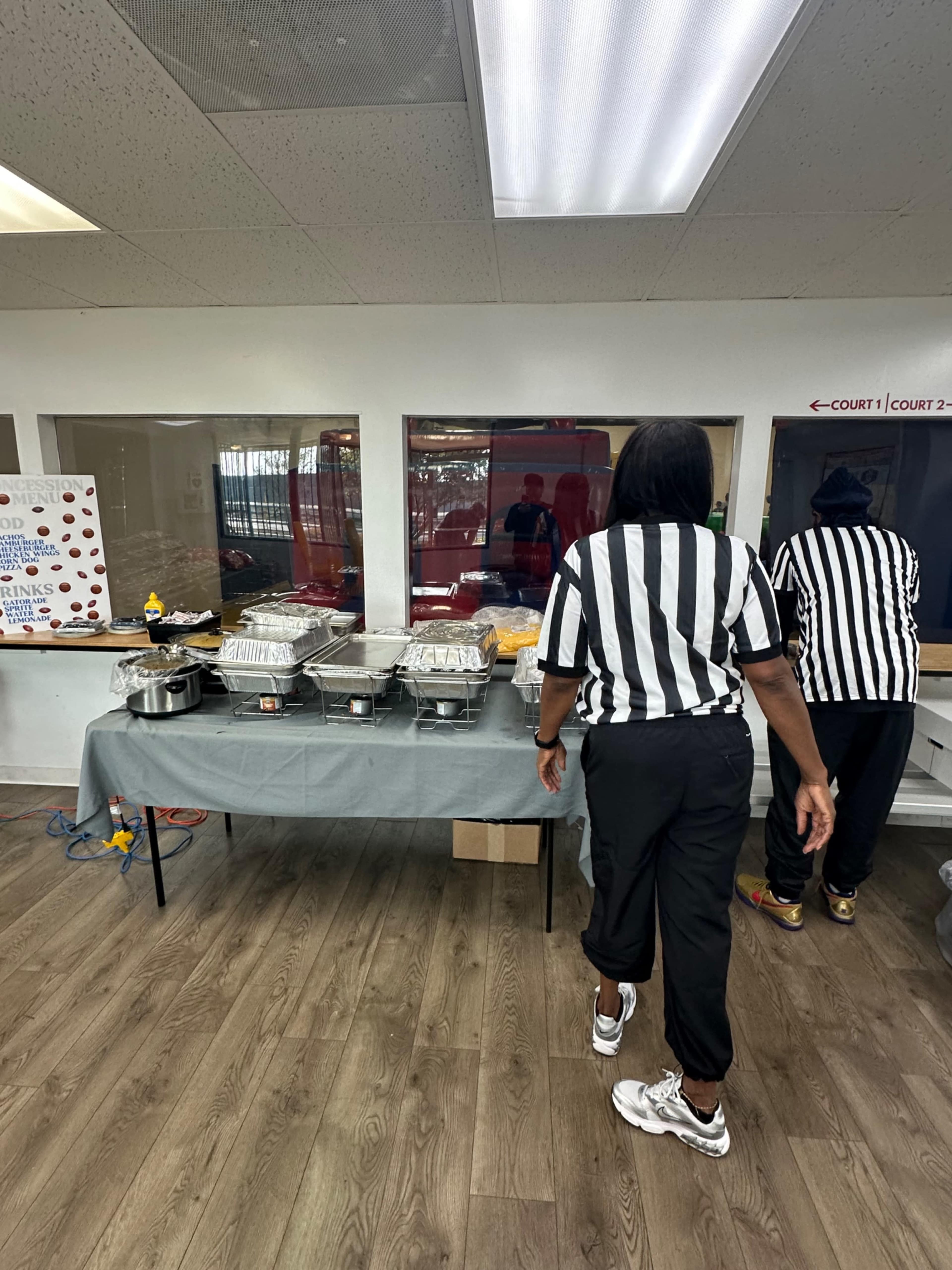 Two individuals wearing referee uniforms are setting up a food table with trays in a room designed for an event or gathering.