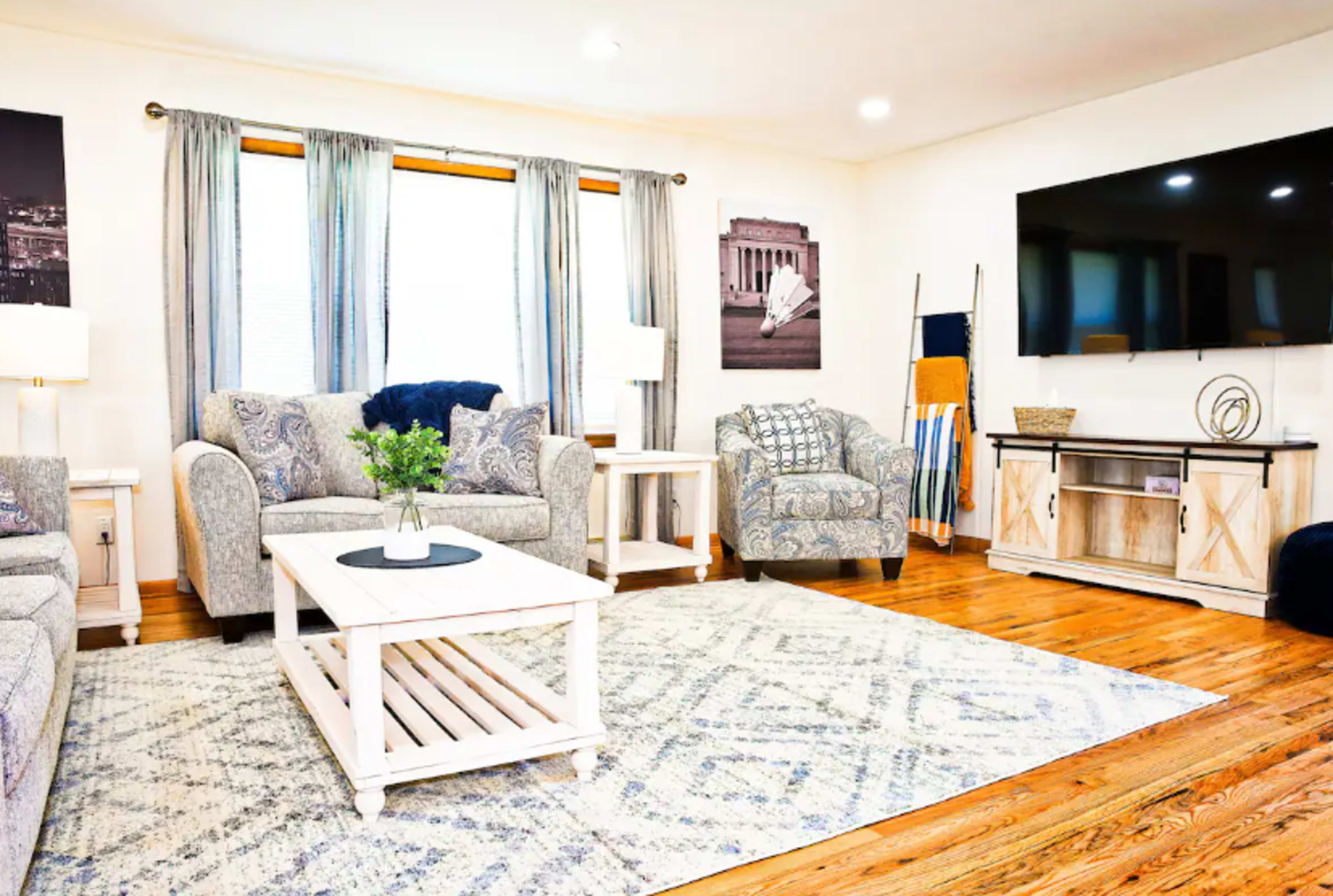 The image shows a well-furnished living room with two patterned sofas, a central coffee table, and a large television mounted on the wall.