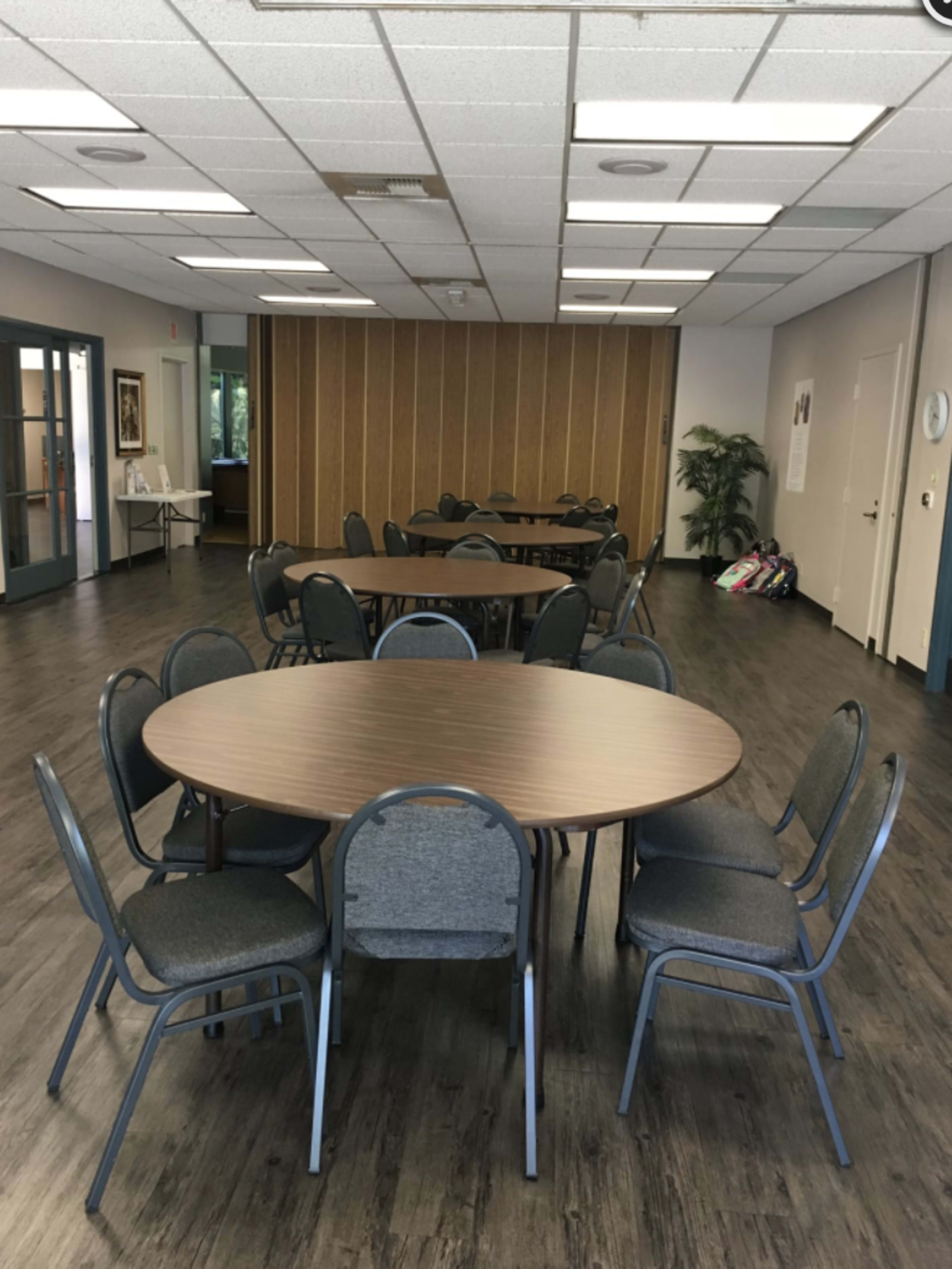 A large meeting room features several round tables surrounded by chairs on a wooden floor, with a wall of wooden panels in the background.