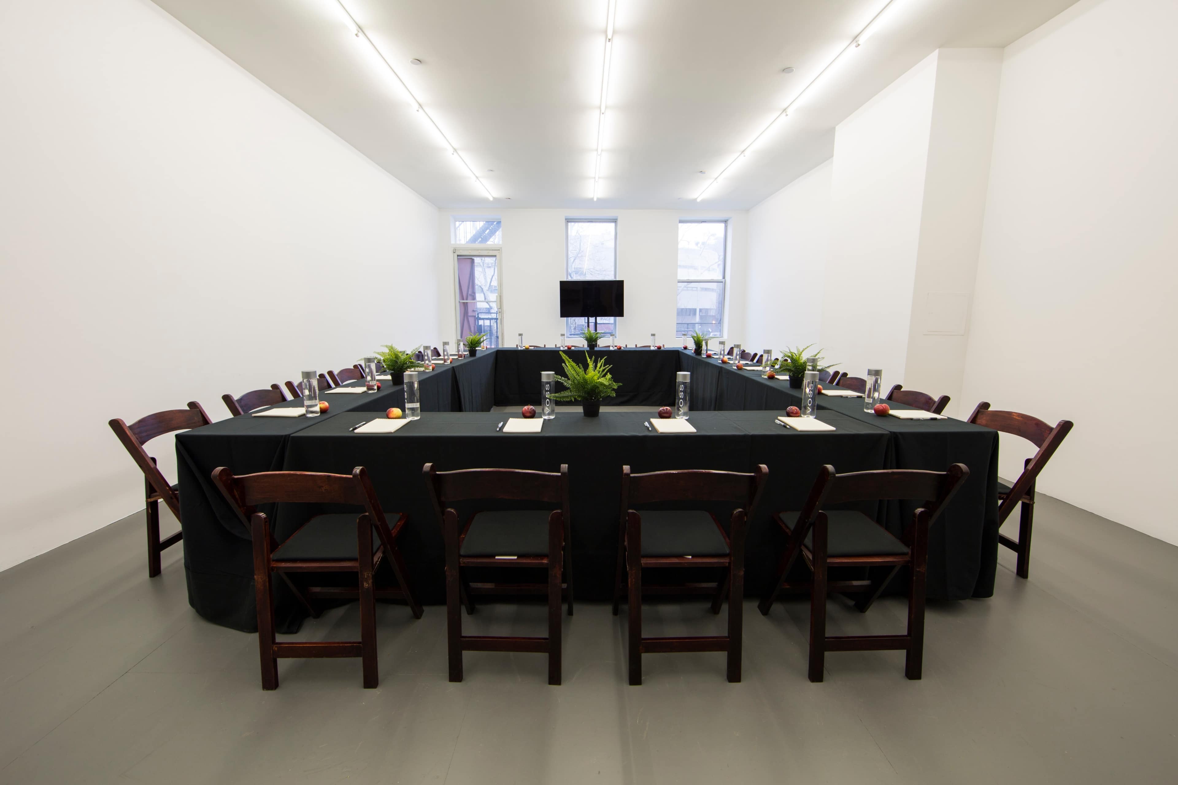 A long table is set up in a spacious, brightly lit room with chairs arranged around it and a television mounted on the wall.