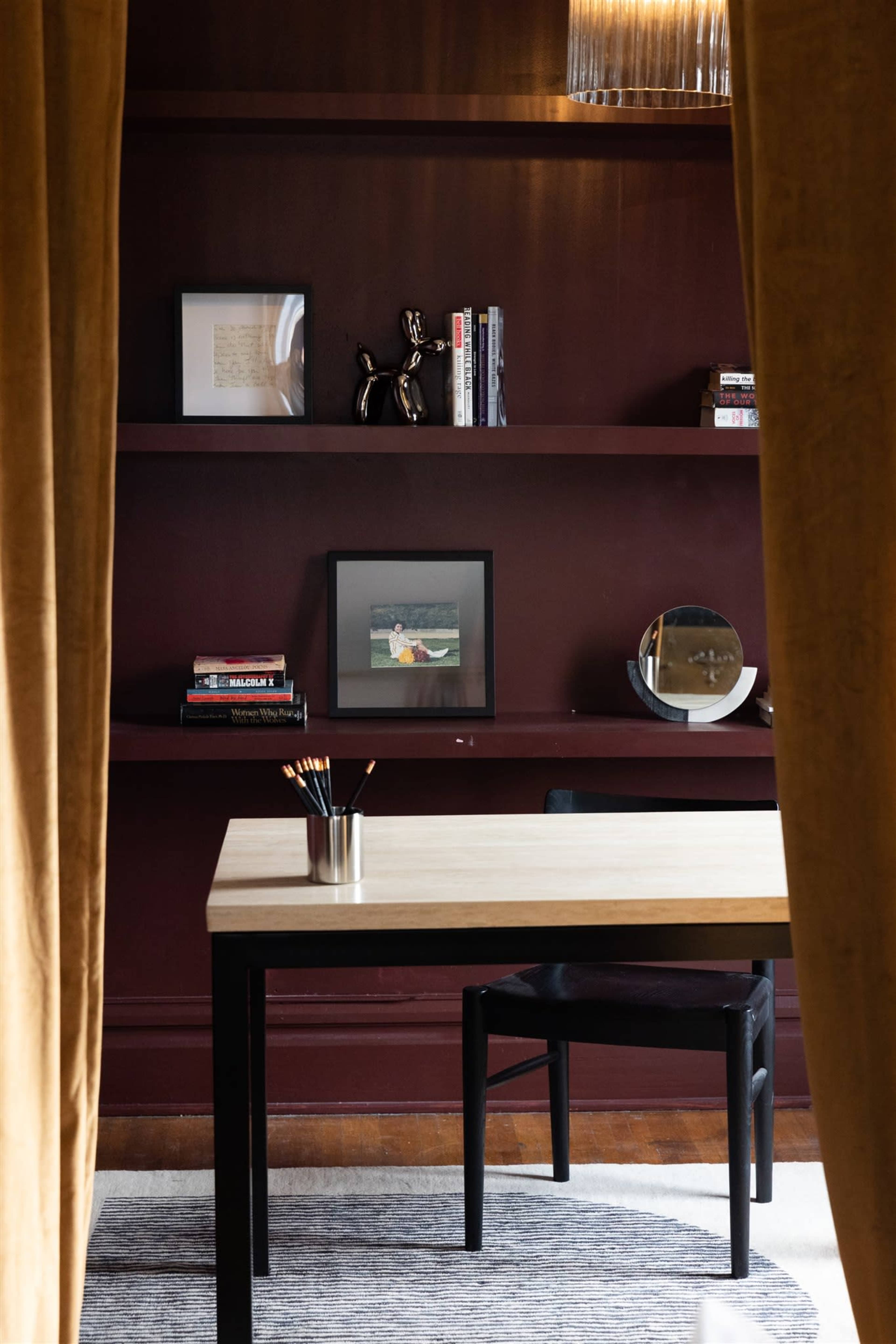 A modern home office features a desk and chair with dark wooden shelves displaying books and framed pictures in a richly colored room.