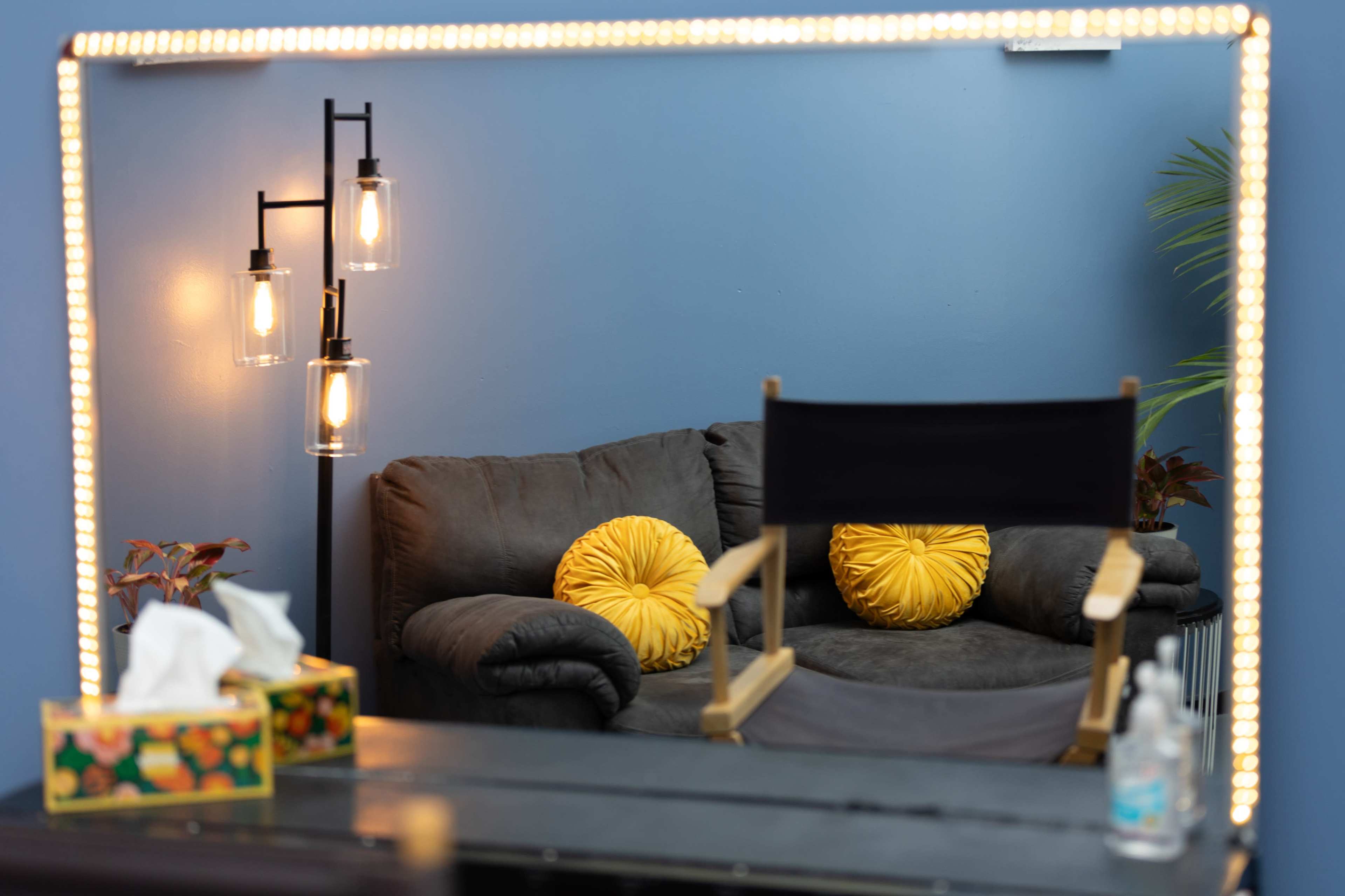A mirror reflecting a cozy seating area with a dark couch, yellow decorative pillows, a standing lamp, and a director's chair in front of a blue wall.