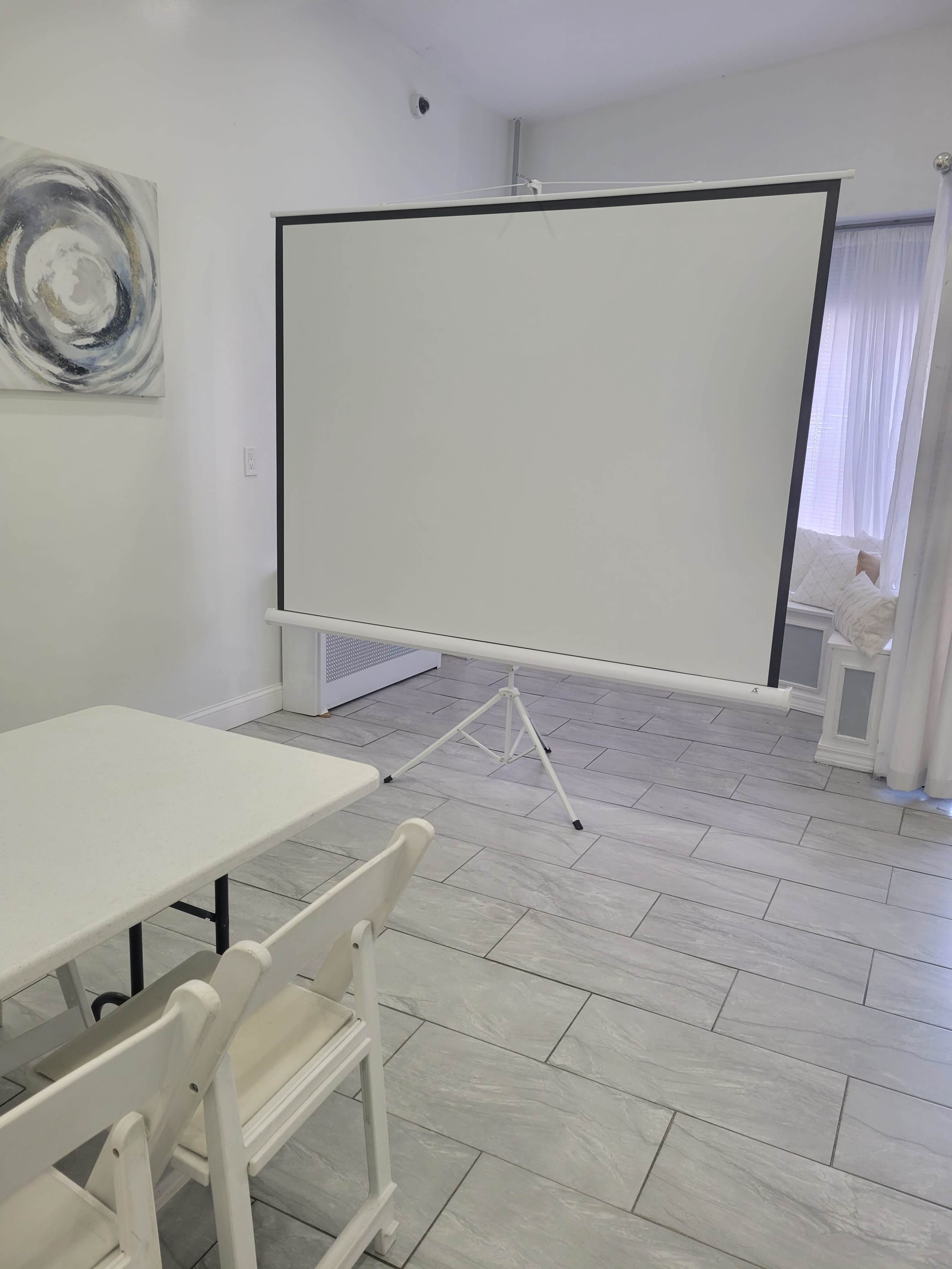 A spacious room featuring a white projector screen on a stand, a small table, and several white chairs arranged in front of it.