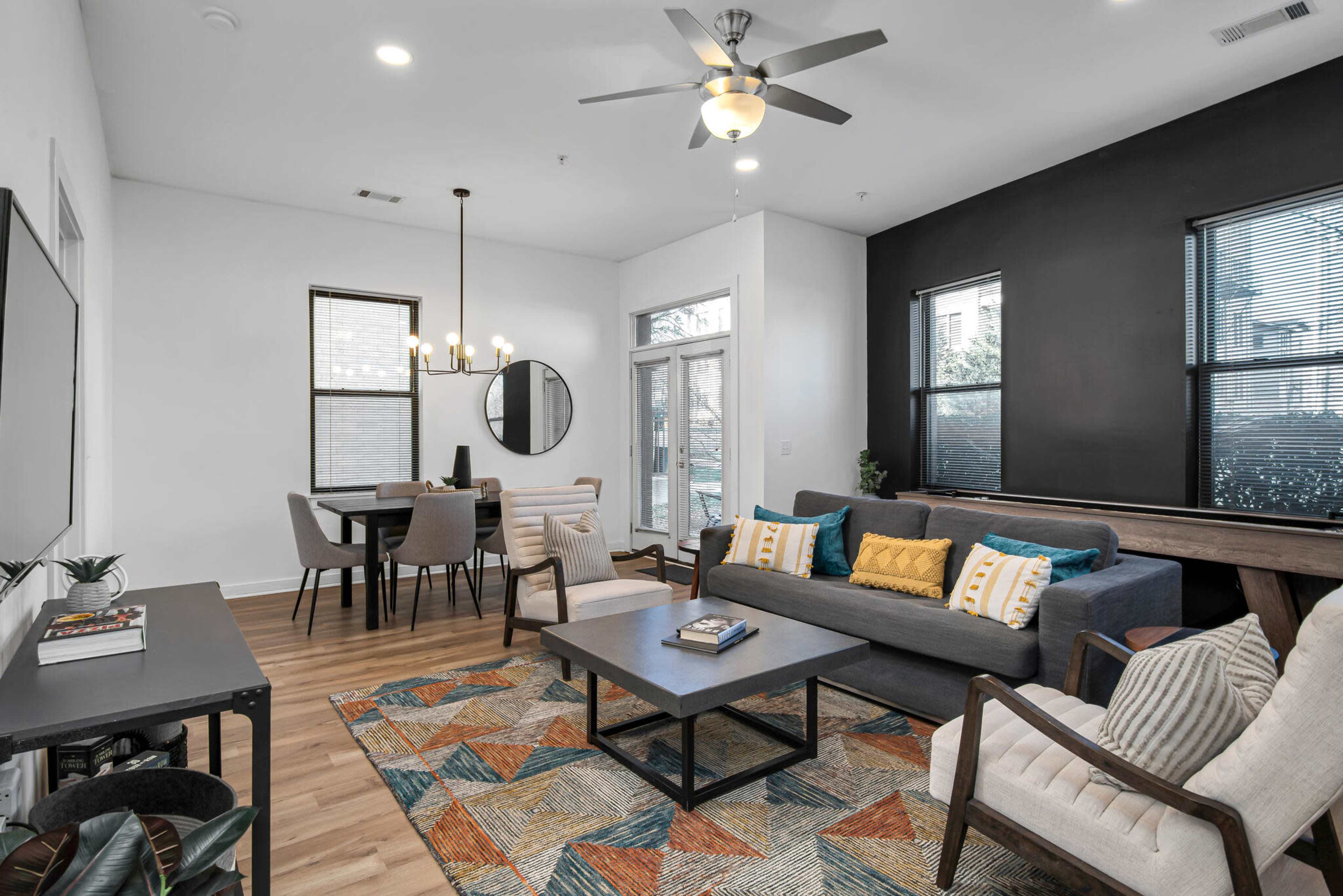 The image shows a modern living room with a sofa, chairs, a coffee table, and a dining area, featuring large windows and a geometric-patterned rug.