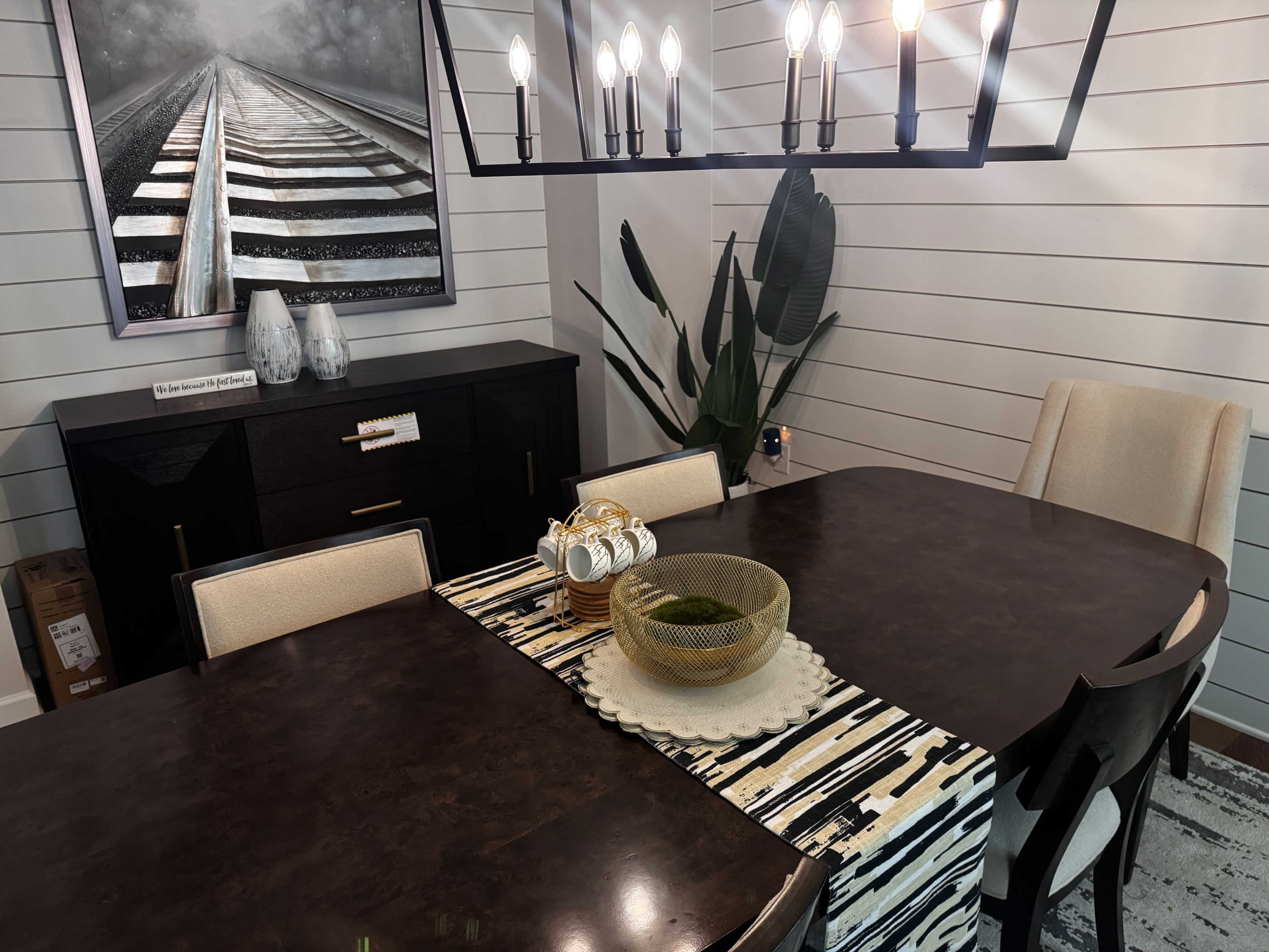A dining room features a dark wooden table surrounded by cream-colored chairs, with a decorative table runner, a bowl of greenery, and a light fixture overhead, alongside a sideboard and wall art depicting train tracks.