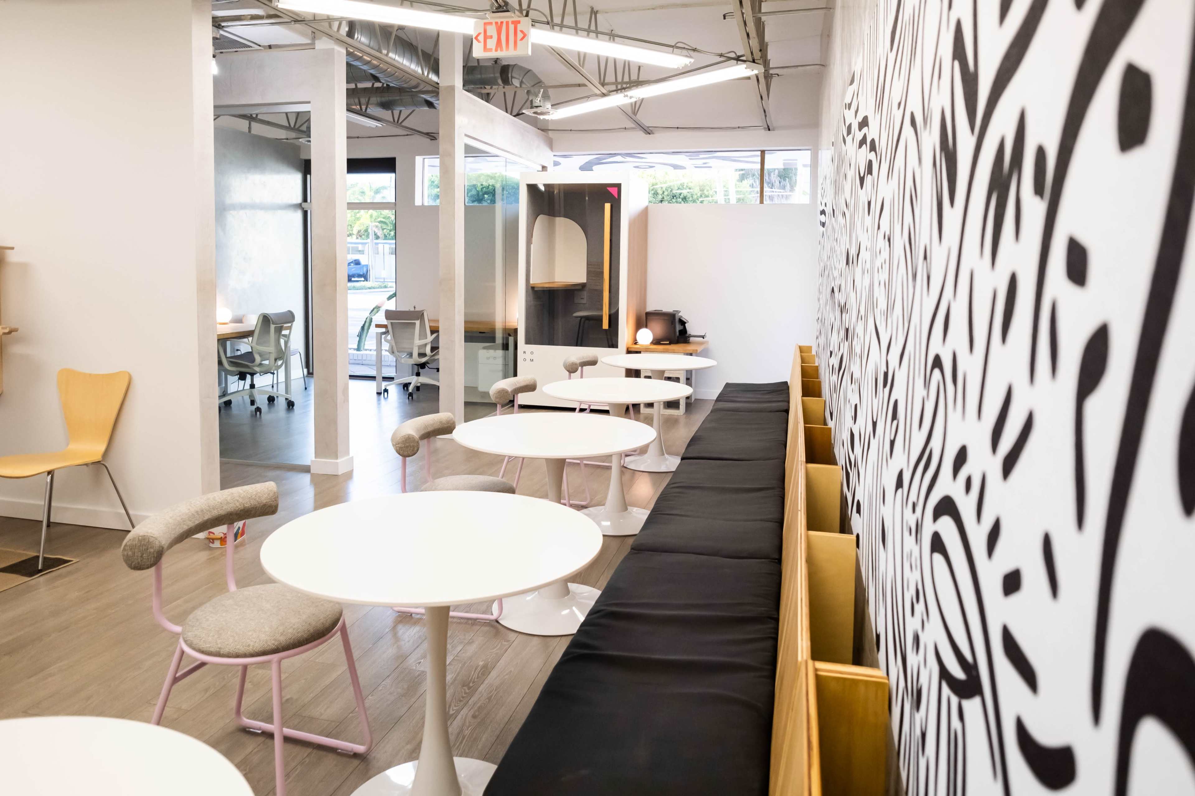 The image shows a modern office space with a long black bench, circular tables, and a patterned wall.