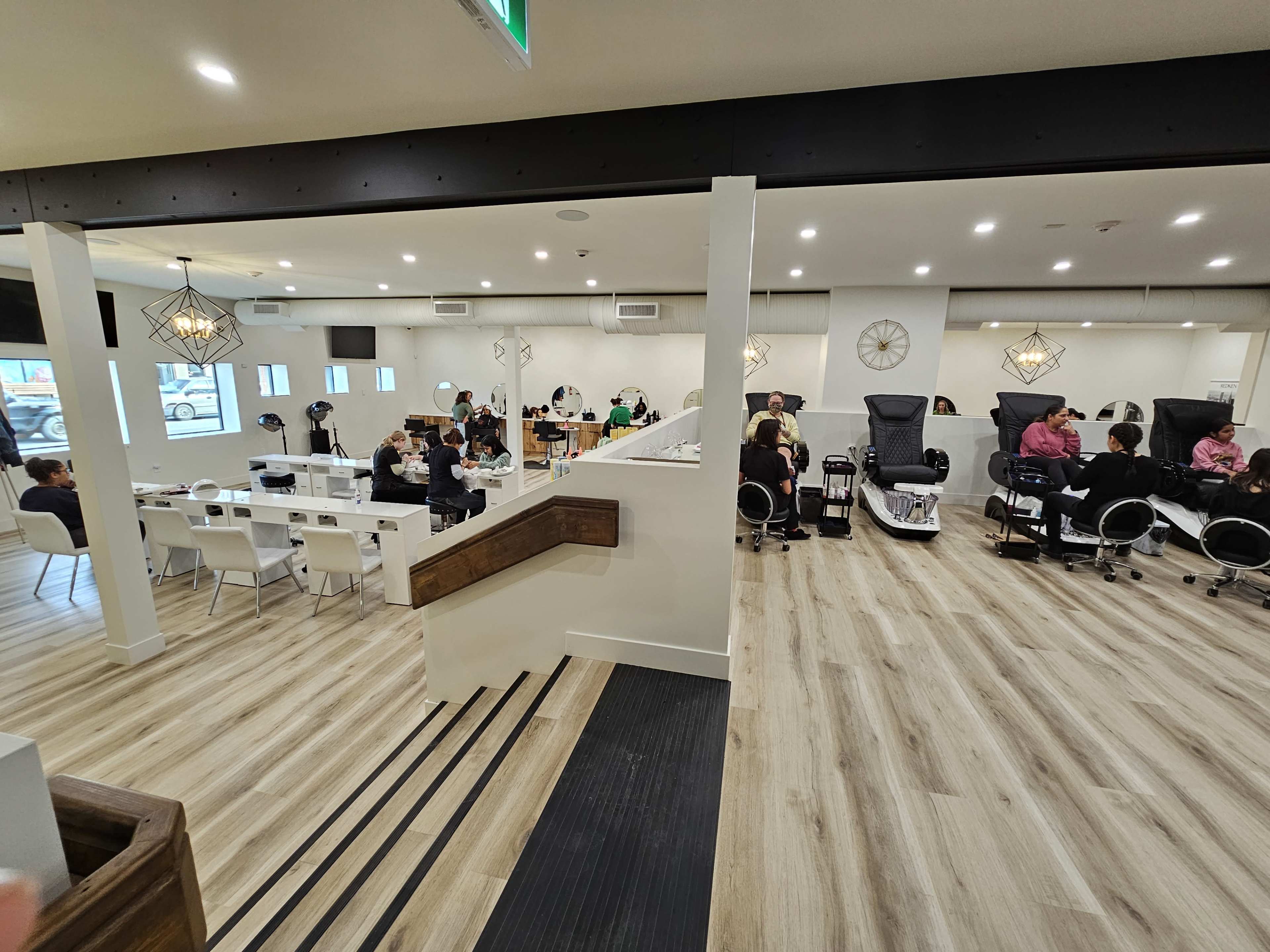 The image shows a well-organized nail salon with several clients receiving manicures and pedicures in an airy, modern setting.