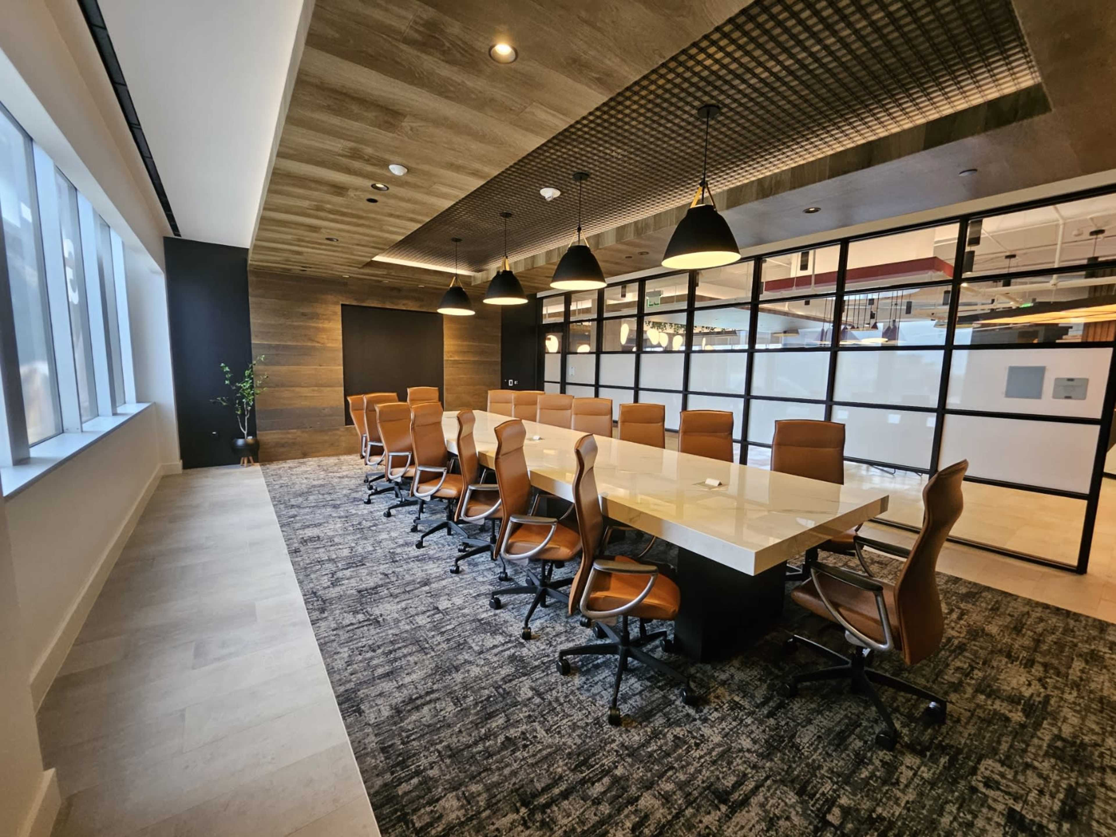 A large conference room features a long, rectangular table surrounded by brown leather chairs, with modern lighting and large windows.