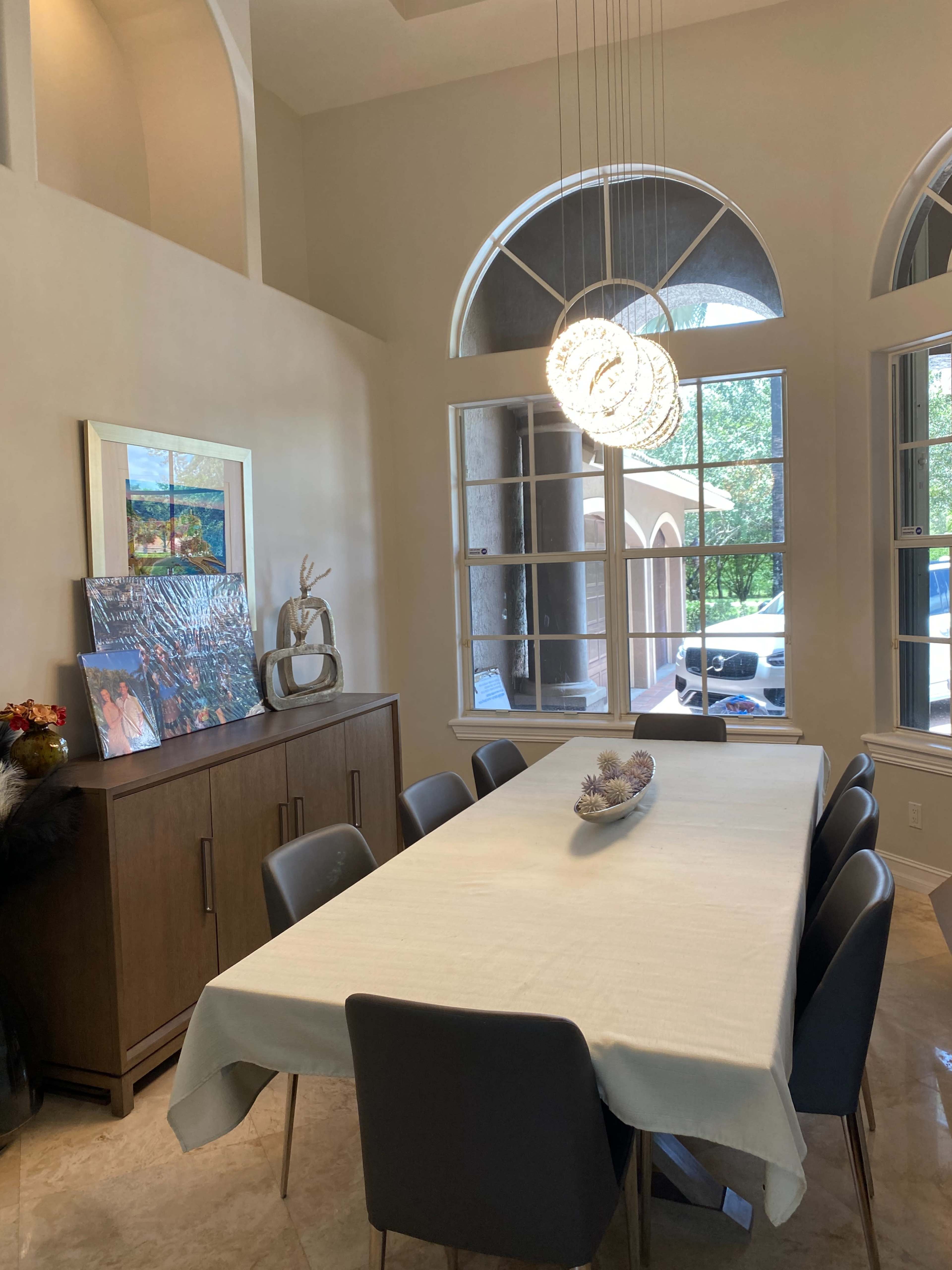 A long dining table is set in a well-lit room, flanked by modern chairs and large windows that allow natural light to enter.
