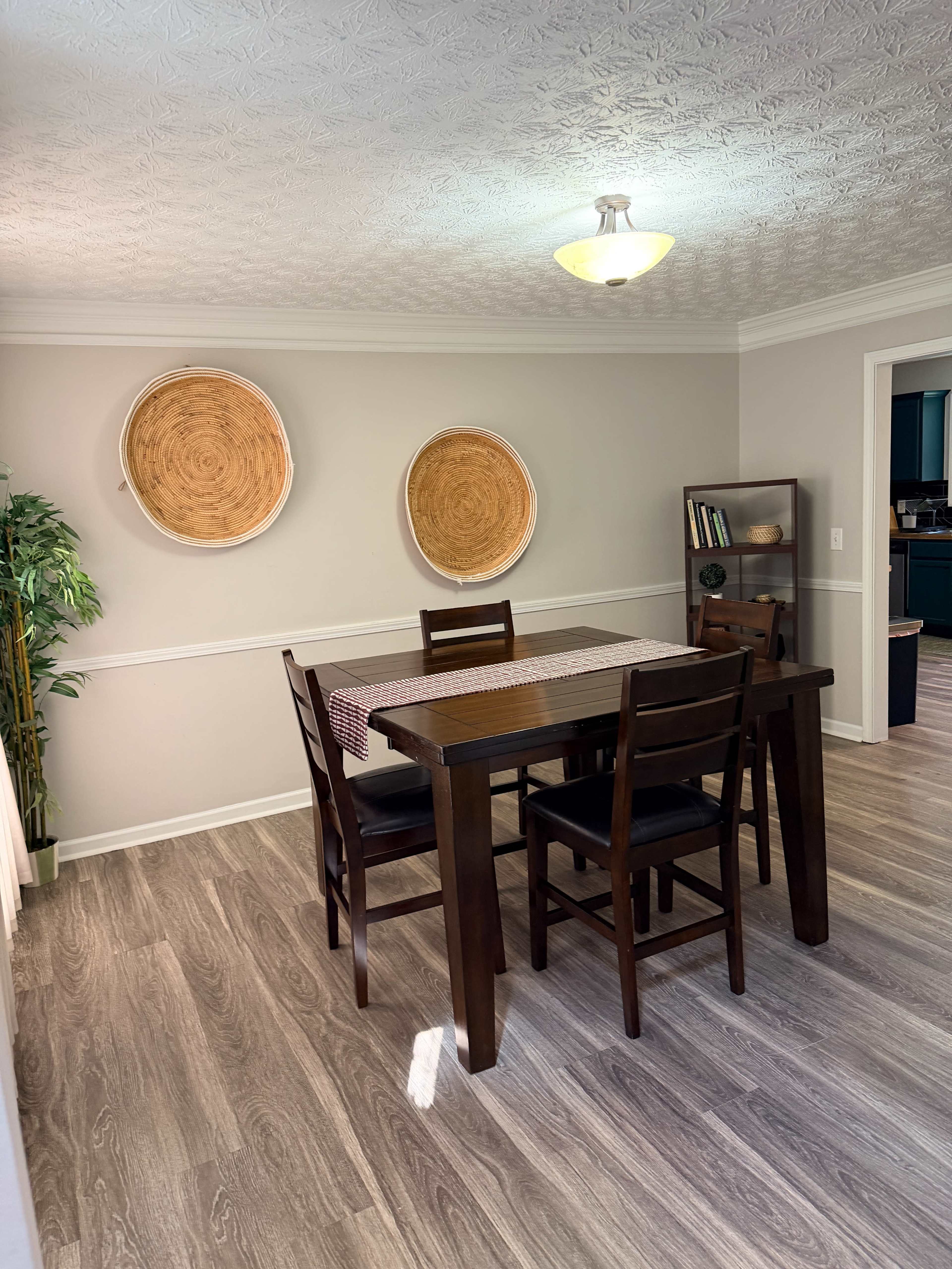 The image shows a dining area with a wooden table and chairs, featuring two decorative wall hangings and a small shelf against the wall.