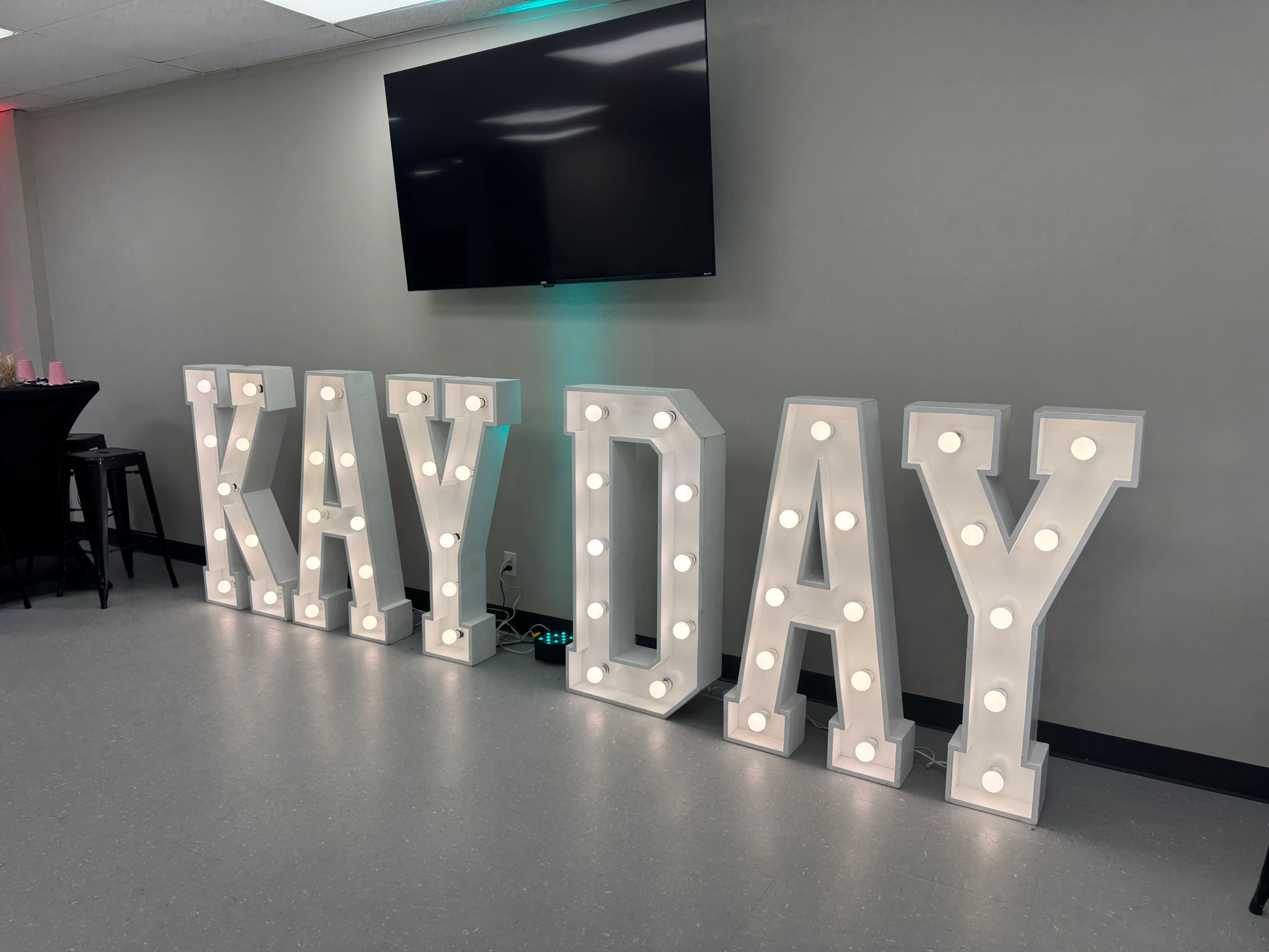 The image shows large illuminated letters spelling out "KAY DAY" against a gray wall with a mounted television above.