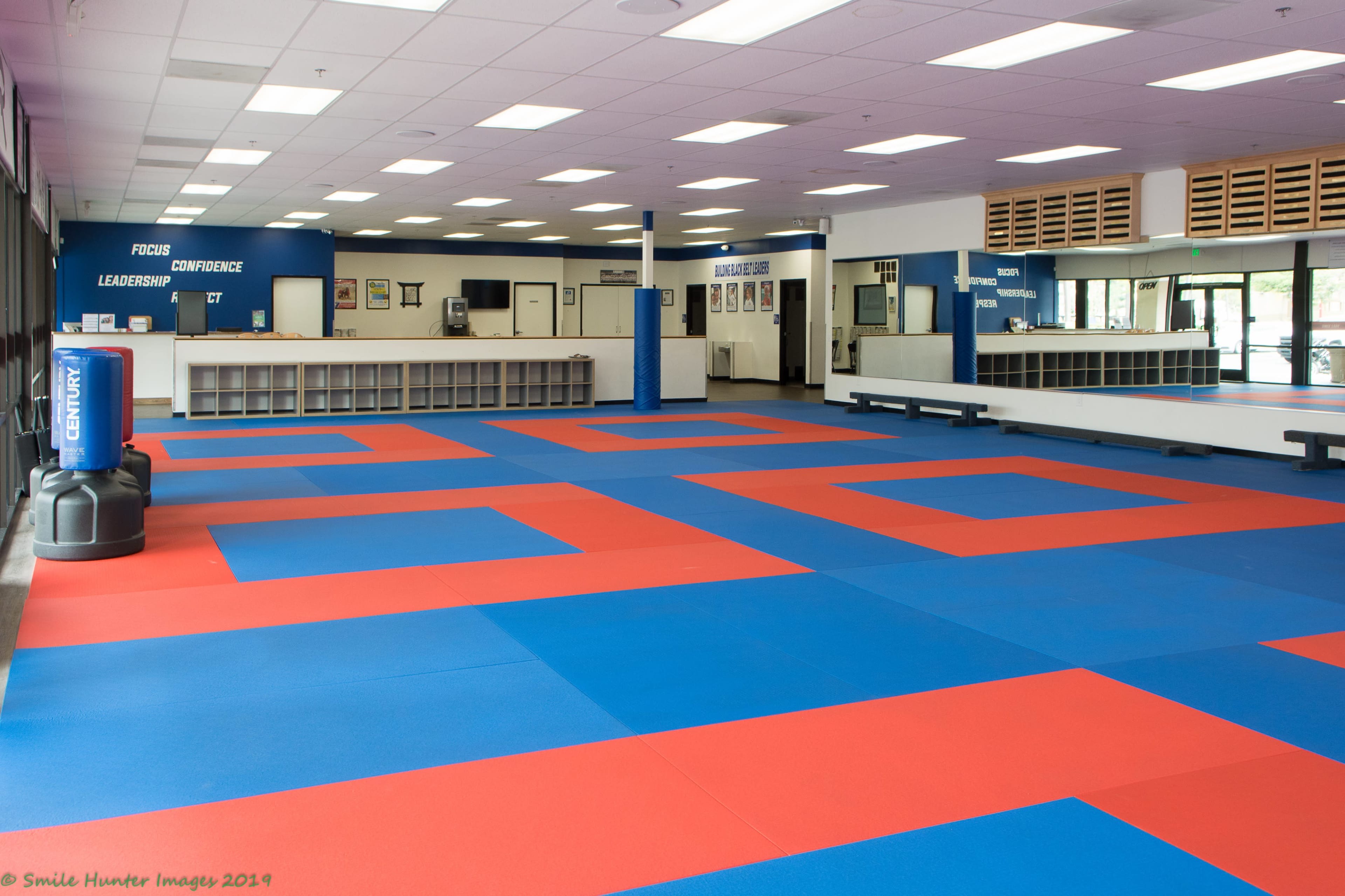A spacious martial arts studio featuring red and blue mats, with a reception area and motivational signs on the walls.