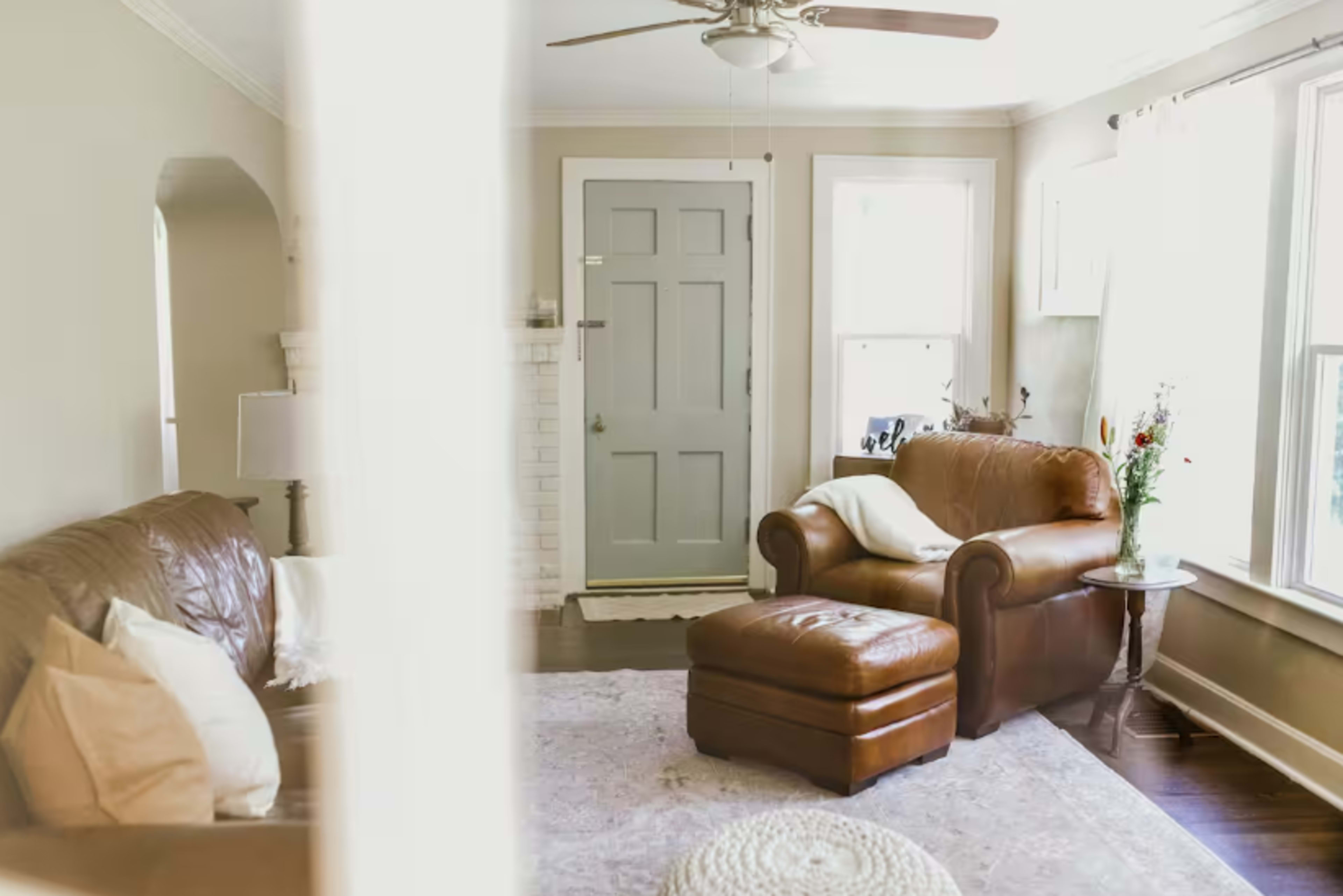 A cozy living room features a brown leather couch, an ottoman, and a light-colored rug, with a blue door in the background and natural light streaming through the windows.